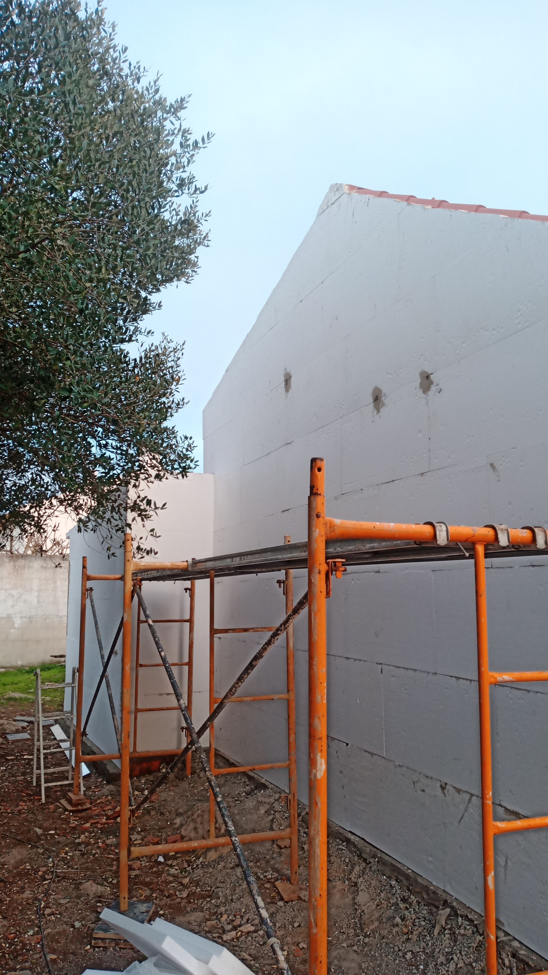 Canteiro de obras: prédio branco com andaimes. Árvore à esquerda, cascalho e terra no chão.