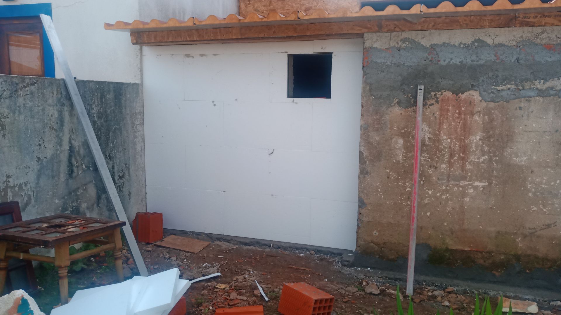 Parede externa em construção com pequena janela, tijolos e ferramentas.