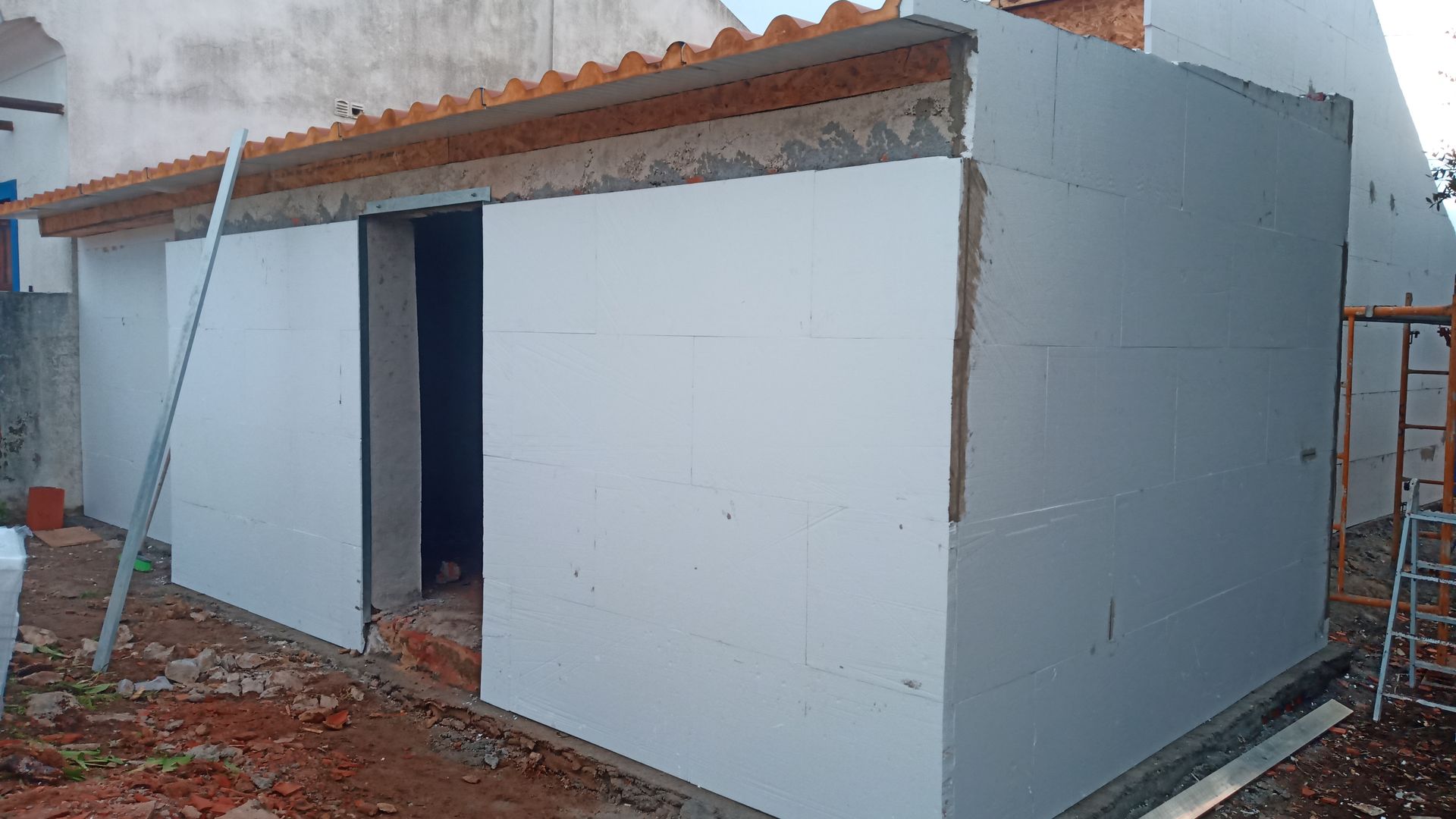 Prédio em construção com painéis isolantes brancos, porta aberta e telhado de telha vermelha.