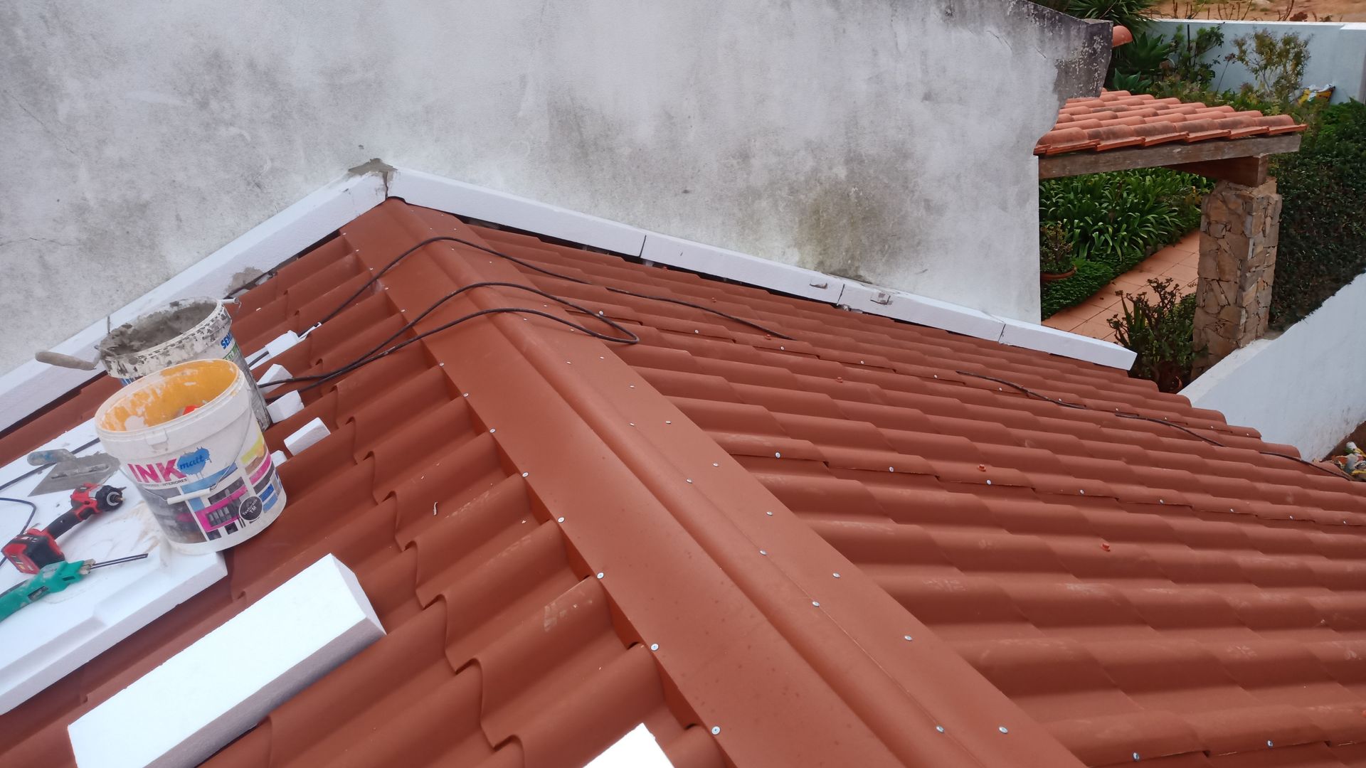 Telhado de telhas vermelhas em obras, com ferramentas e materiais visíveis. Parede branca ao fundo.