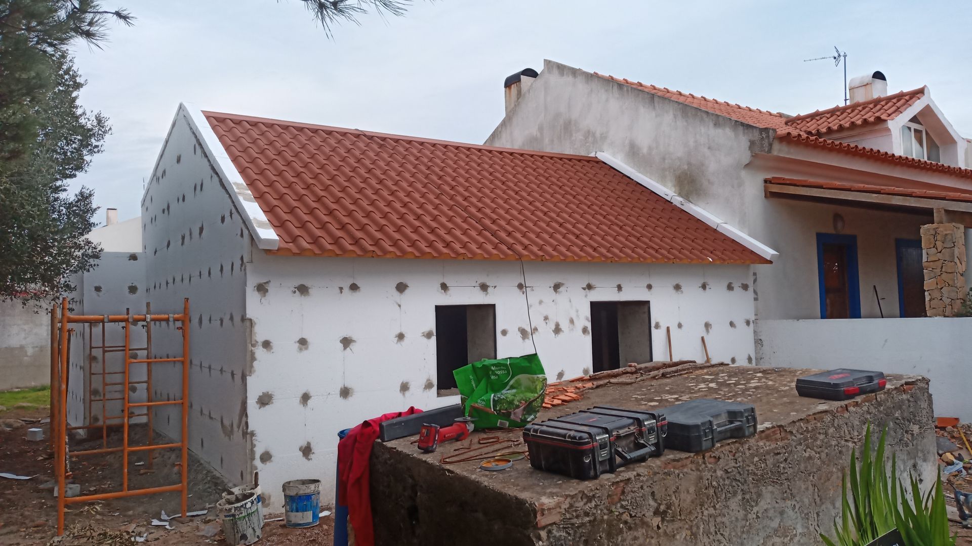 Exterior de prédio em construção com telhado de telha vermelha, paredes brancas com isolamento e ferramentas no quintal.