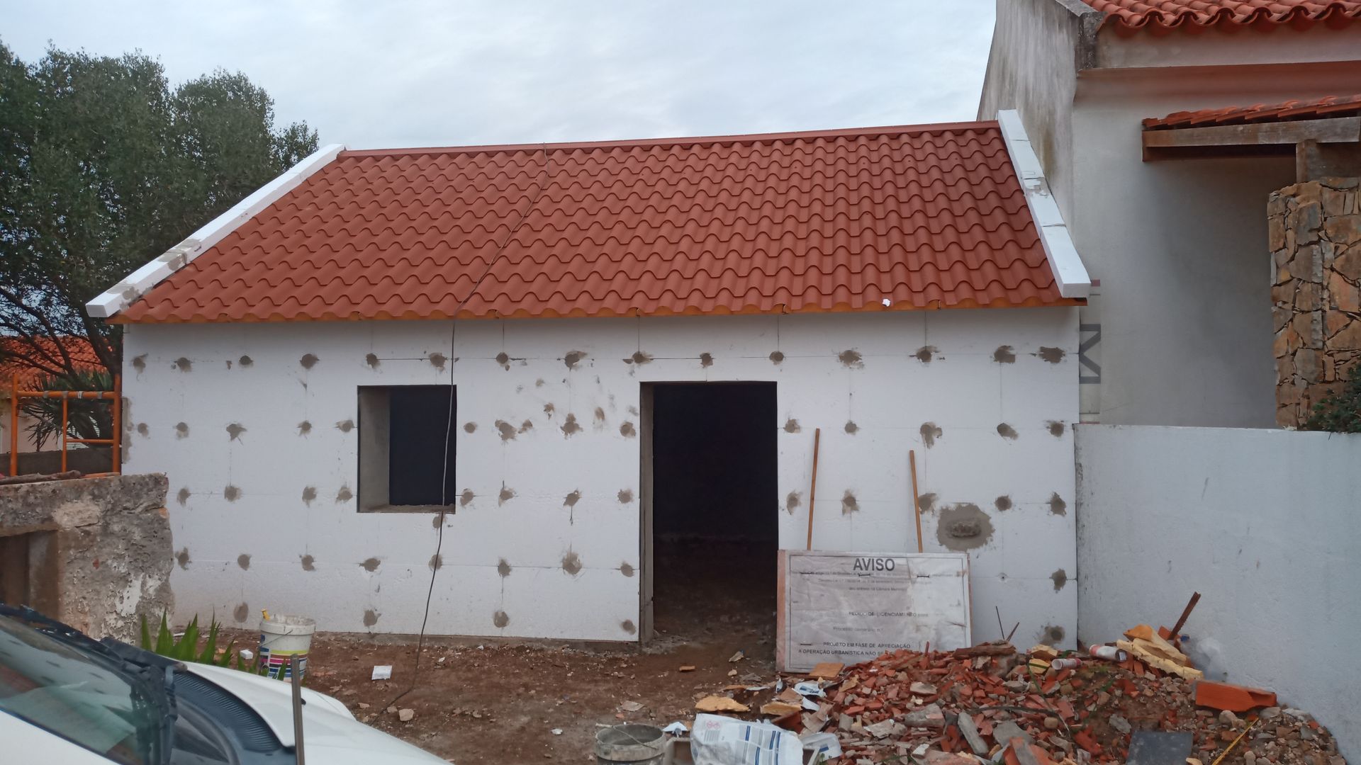 Pequeno prédio branco em construção com telhado de telha vermelha, porta aberta e janela.