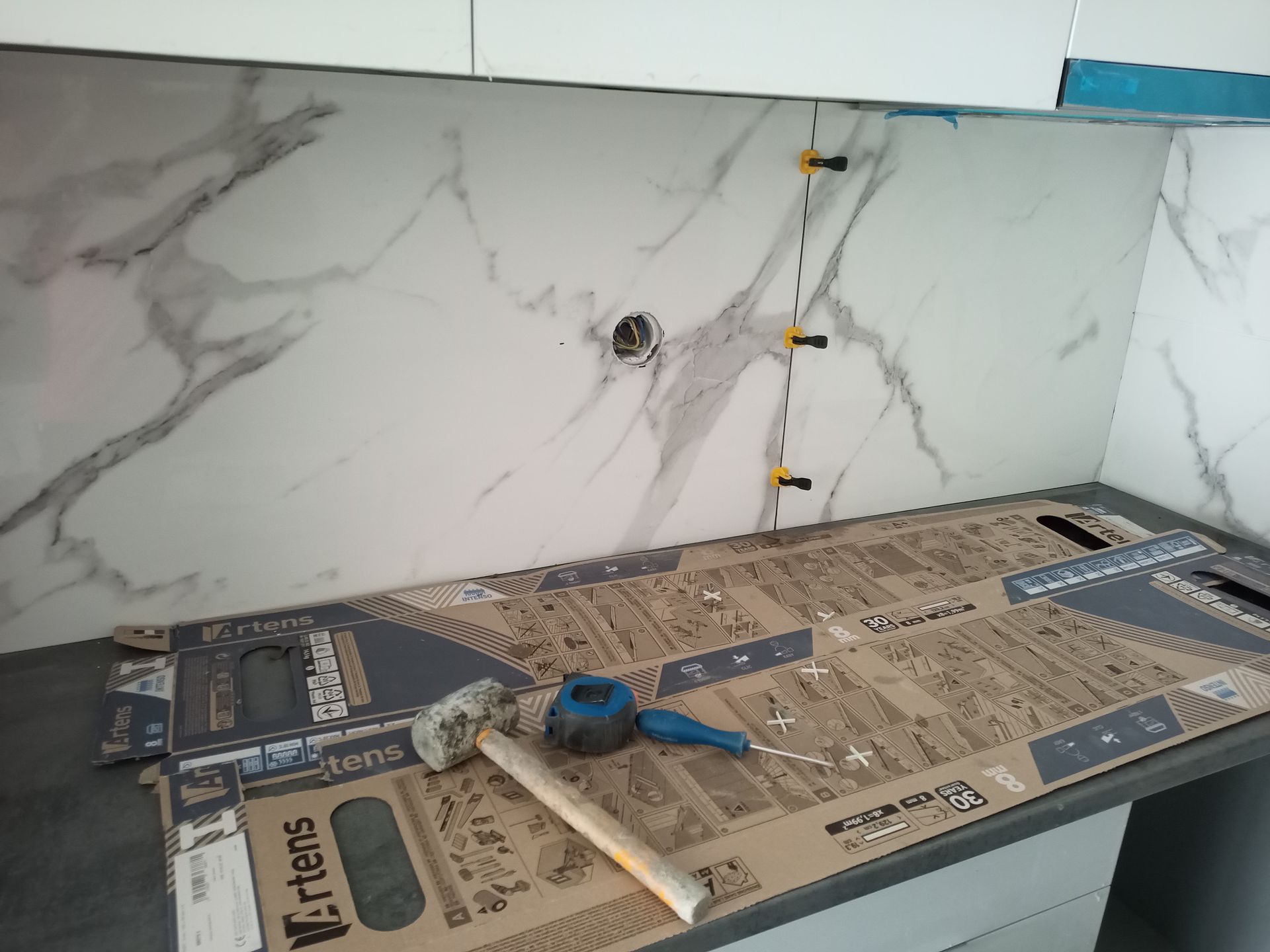 Instalação de azulejos na parede da pia da cozinha. Azulejos com estampa de mármore estão sendo assentados com espaçadores e ferramentas.
