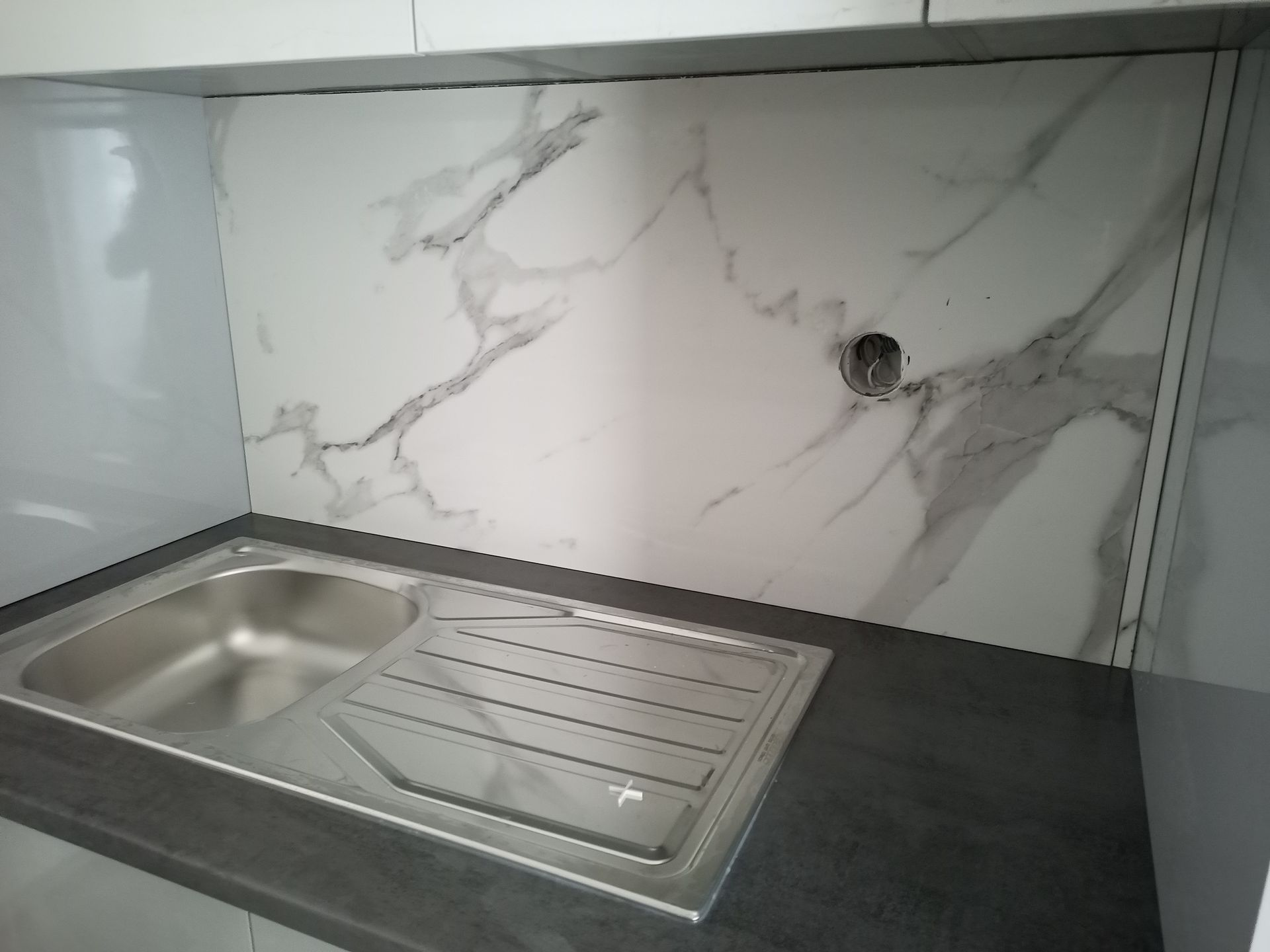Uma pia de cozinha com bancada cinza e backsplash com estampa de mármore; um buraco na parede acima da pia.