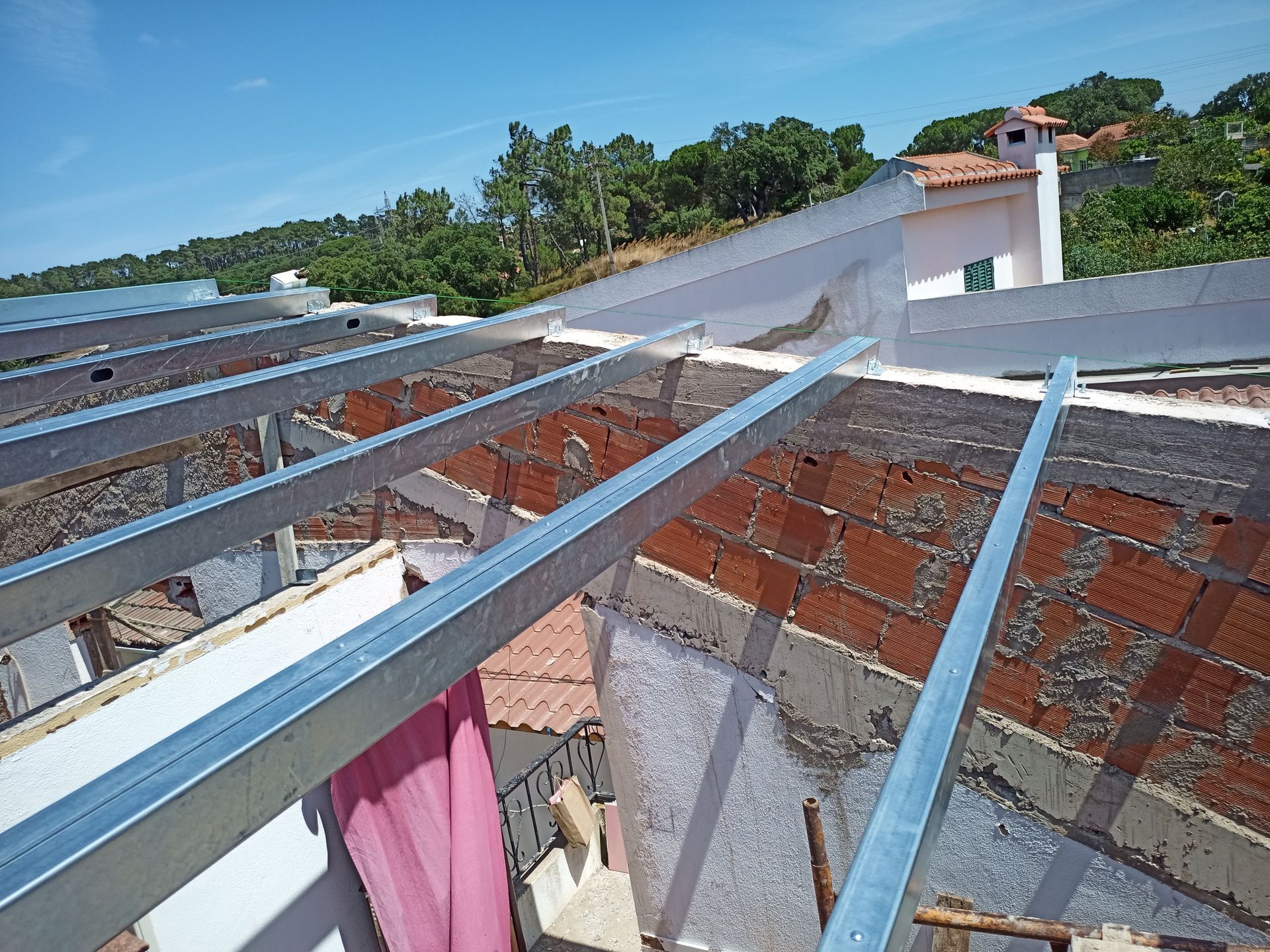 Vigas metálicas dispostas transversalmente sobre uma estrutura de tijolos, vista de cima, provavelmente para um telhado. Céu claro com árvores ao fundo.