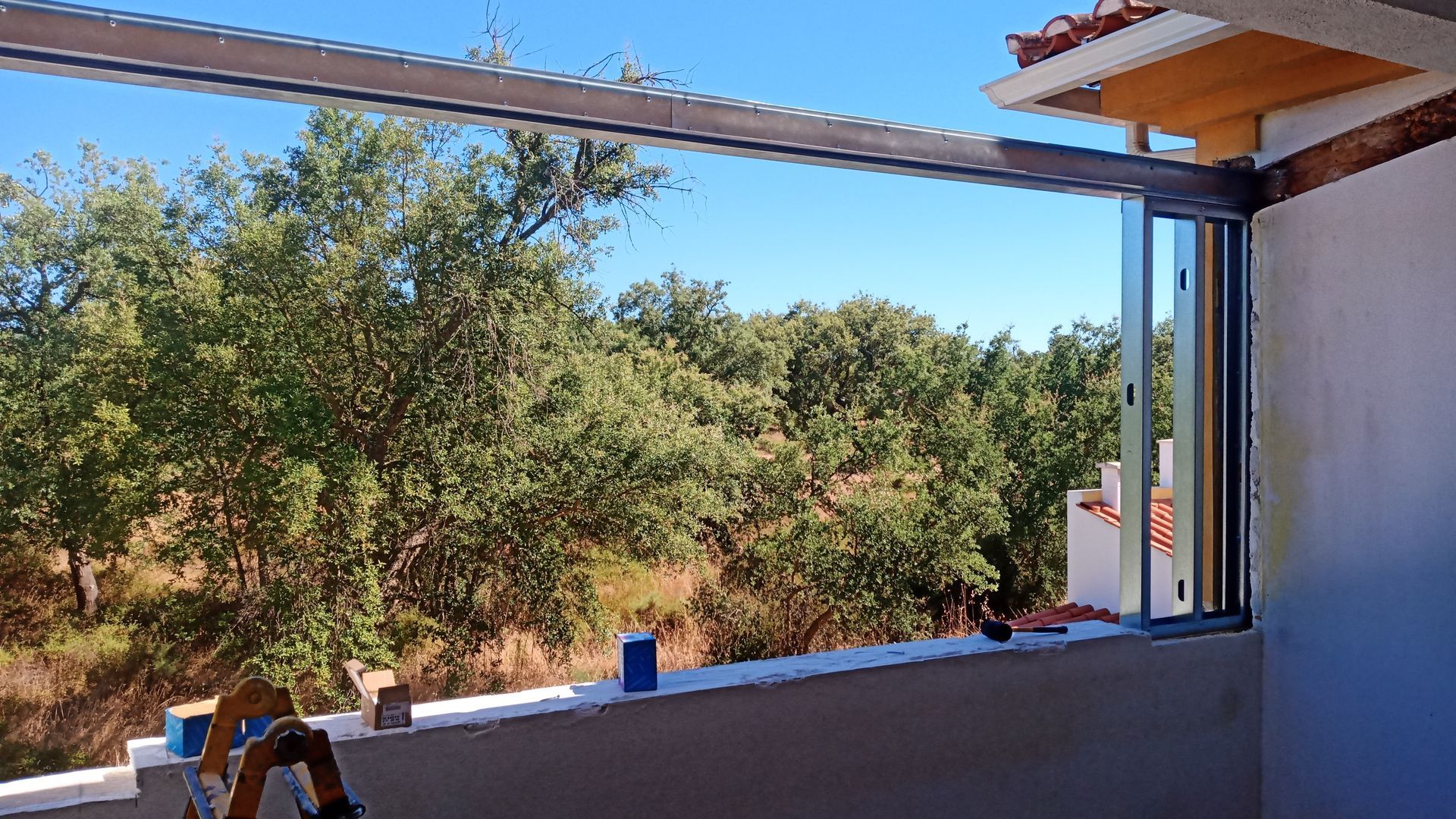 Construção de uma varanda com portas de vidro deslizantes, vigas de aço e vista para árvores verdes sob um céu azul.