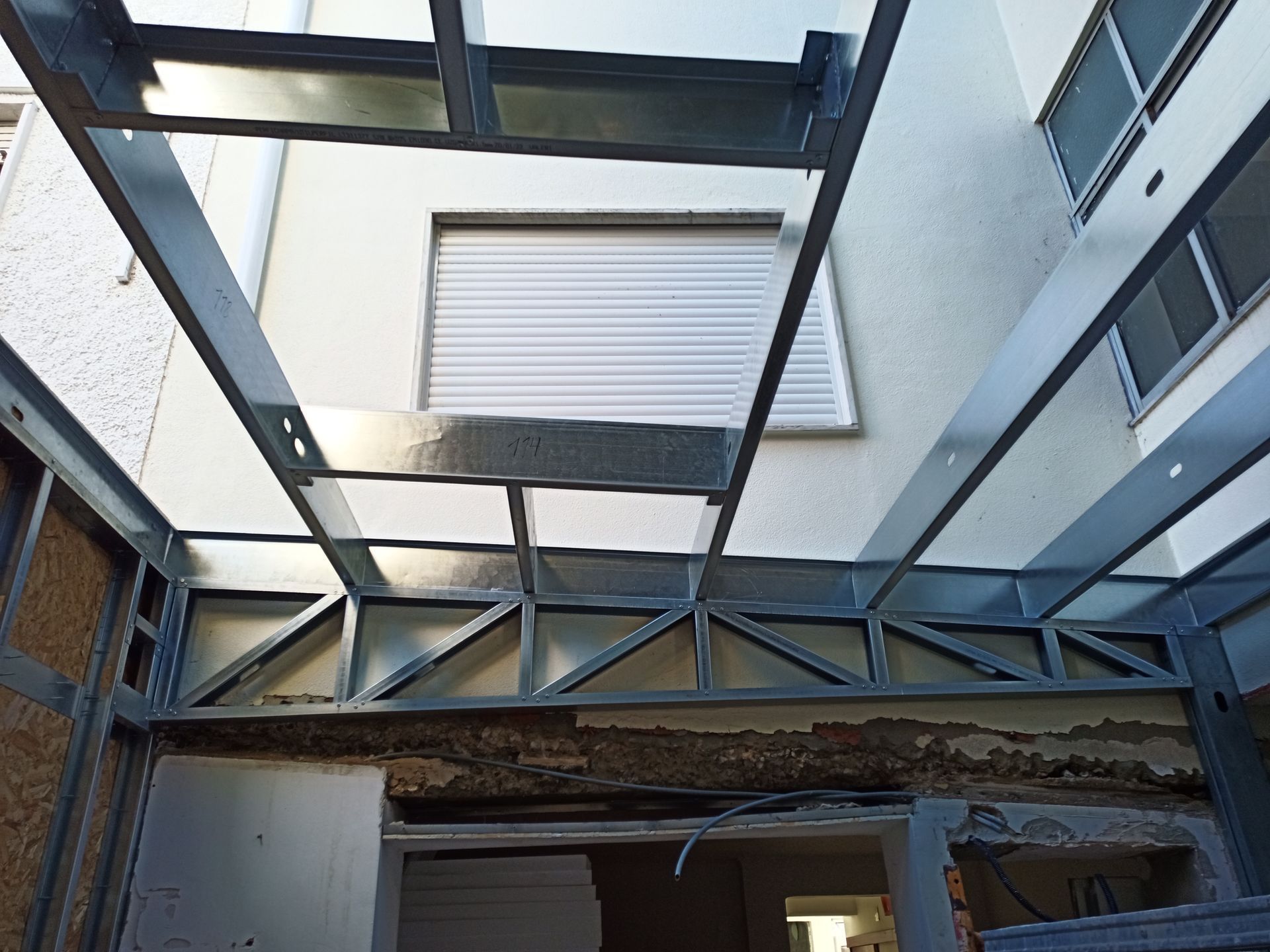 Estrutura metálica de uma claraboia em construção, com uma janela e paredes do edifício ao fundo.