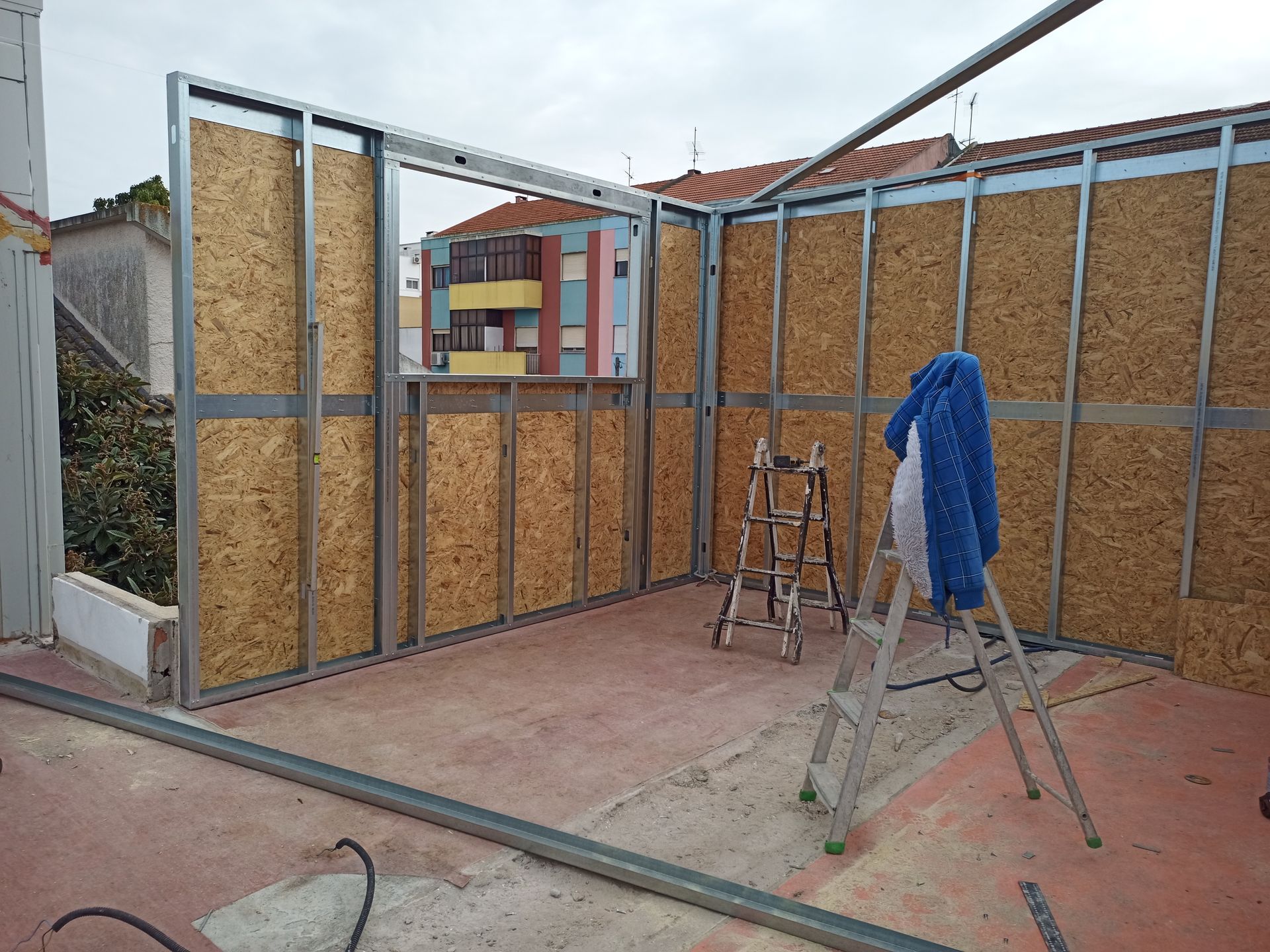 Construção de um pequeno cômodo com estrutura metálica, placas de OSB e uma abertura para janela sobre uma superfície de concreto.