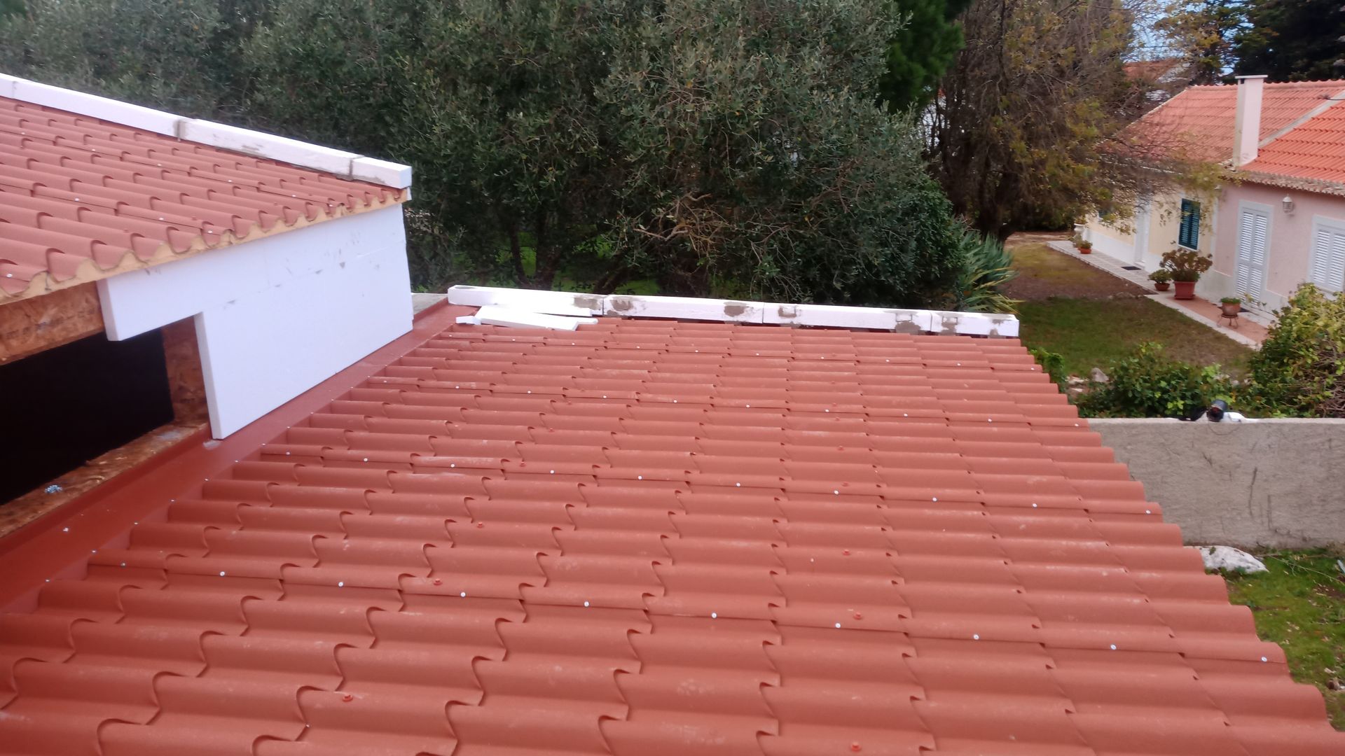 Telhado de telha vermelha com detalhes em branco; árvores e casa ao fundo.