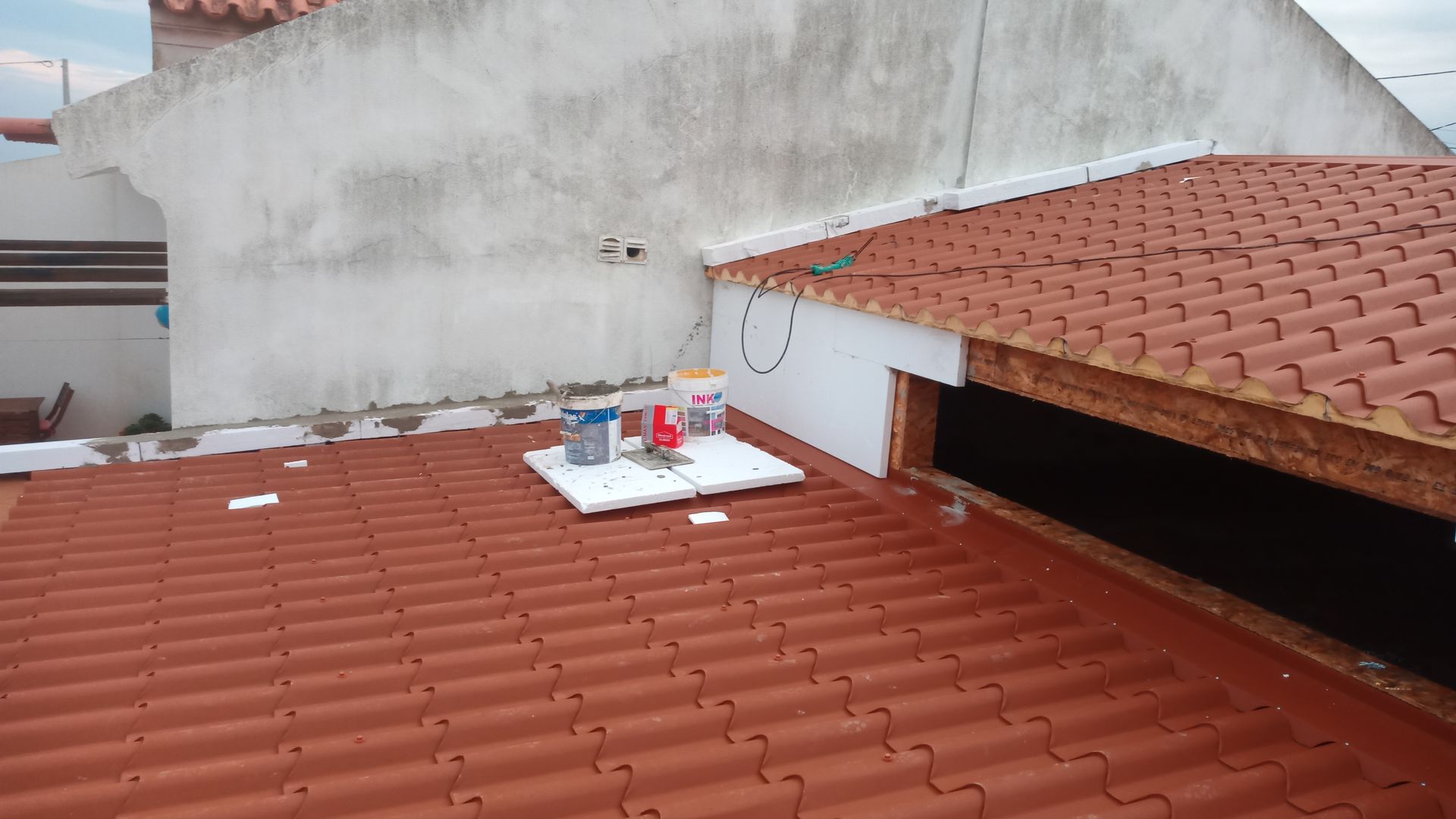 Telhado de telha vermelha com ferramentas, espuma branca e vista parcial de uma parede branca.