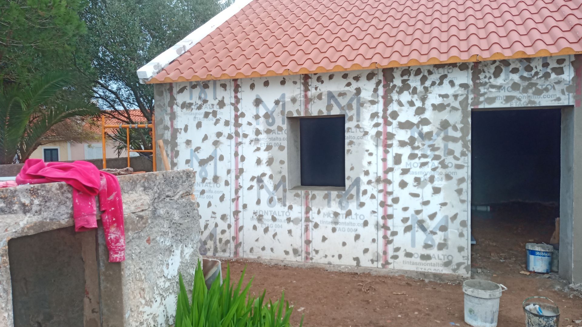 Fachada de prédio em construção com parede aparente, telhado vermelho, janela e porta.
