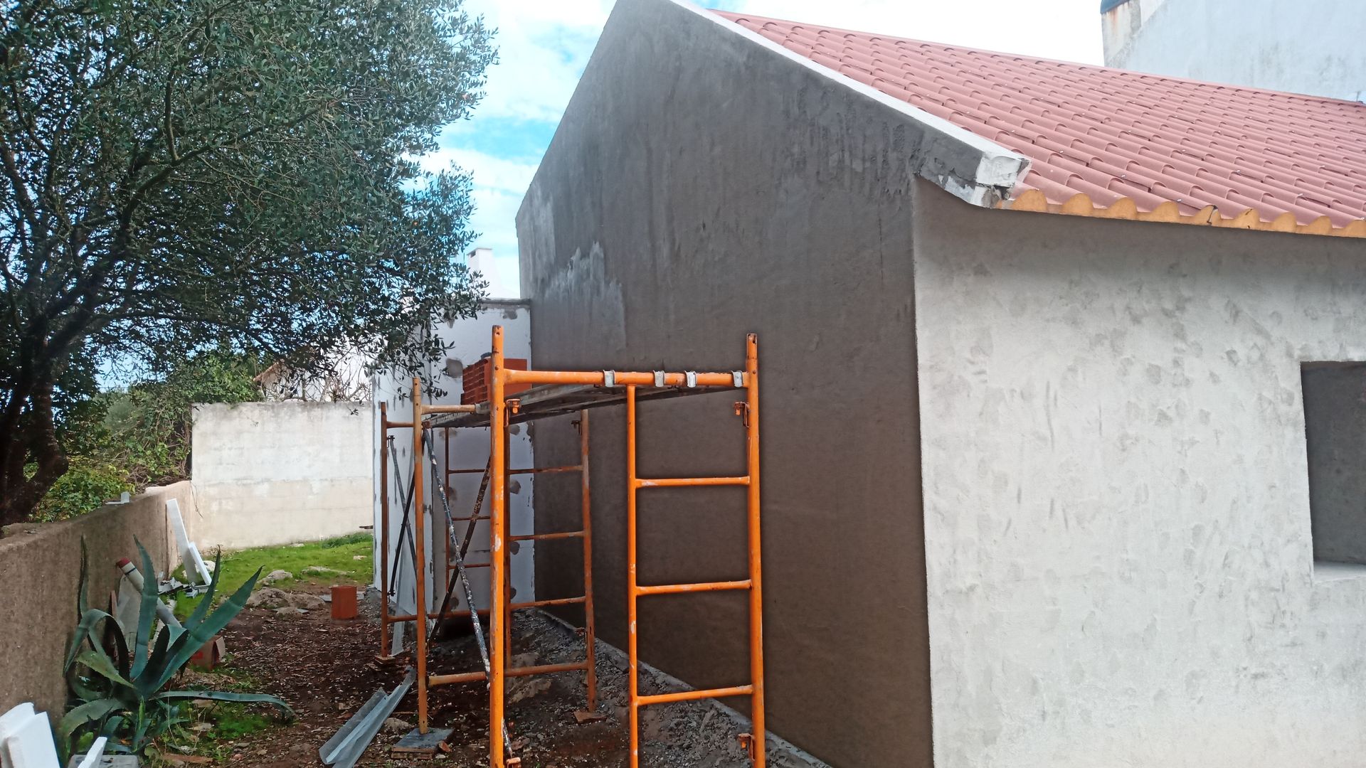 Exterior de um prédio em construção. Estuque marrom na lateral. Andaimes laranja. Telhado de telha vermelha.