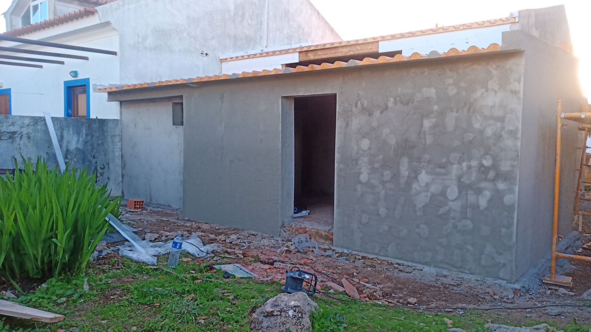 Vista exterior de um edifício em construção, paredes cinzentas, porta aberta, entulho no chão, plantas verdes em primeiro plano.