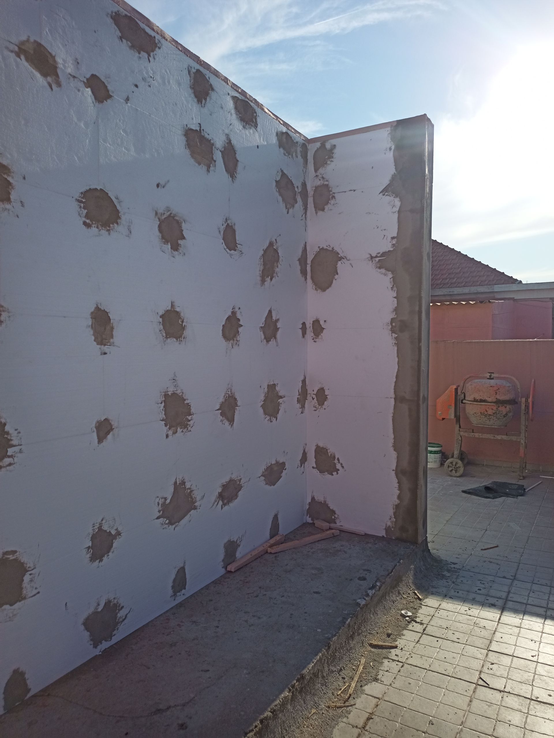 Canto de uma parede branca com manchas marrons espalhadas, em um ambiente externo com piso de azulejo e céu azul.