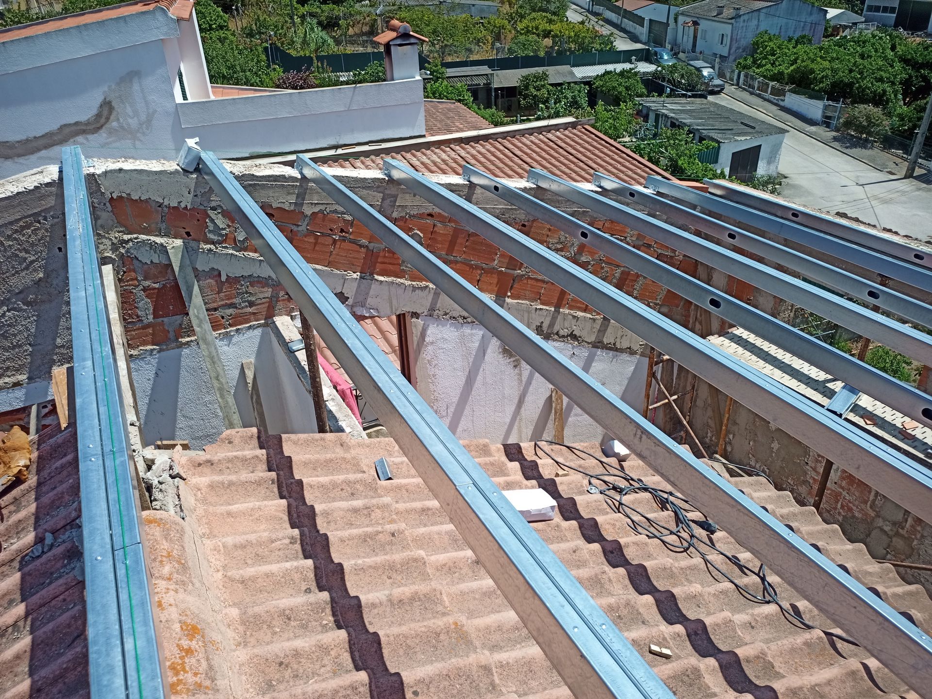 Vigas metálicas estão dispostas transversalmente sobre um telhado de telhas vermelhas, provavelmente para a instalação de painéis solares.