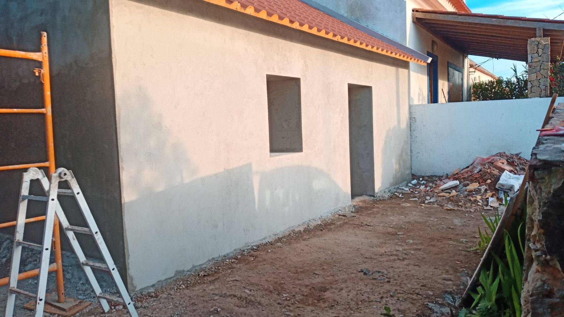 Prédio em construção; parede rebocada cinza com duas aberturas, chão de terra batida, escada à esquerda.