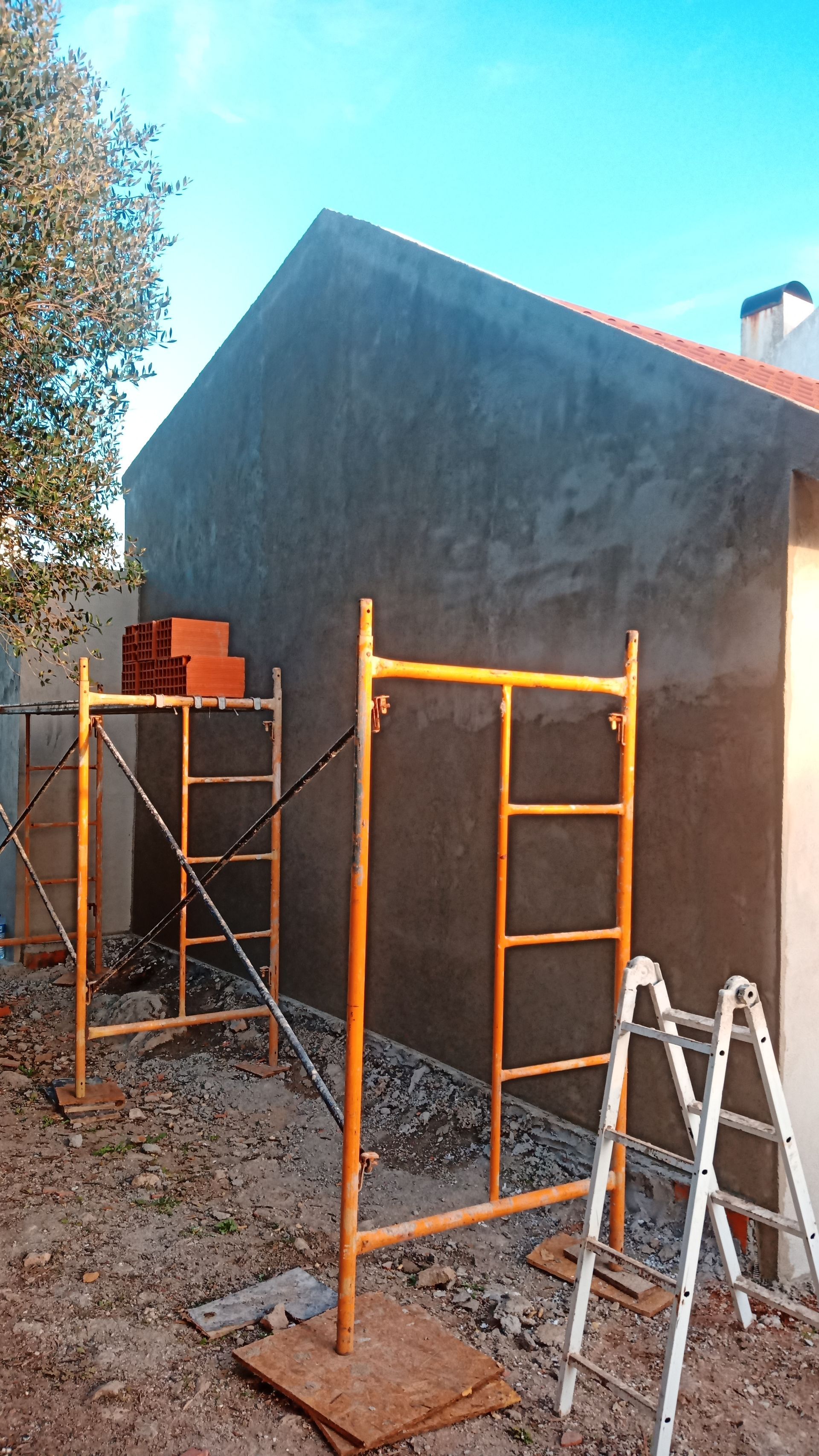 Fachada de prédio em construção; andaimes, tijolos e escada visíveis. Parede de estuque cinza, céu azul.