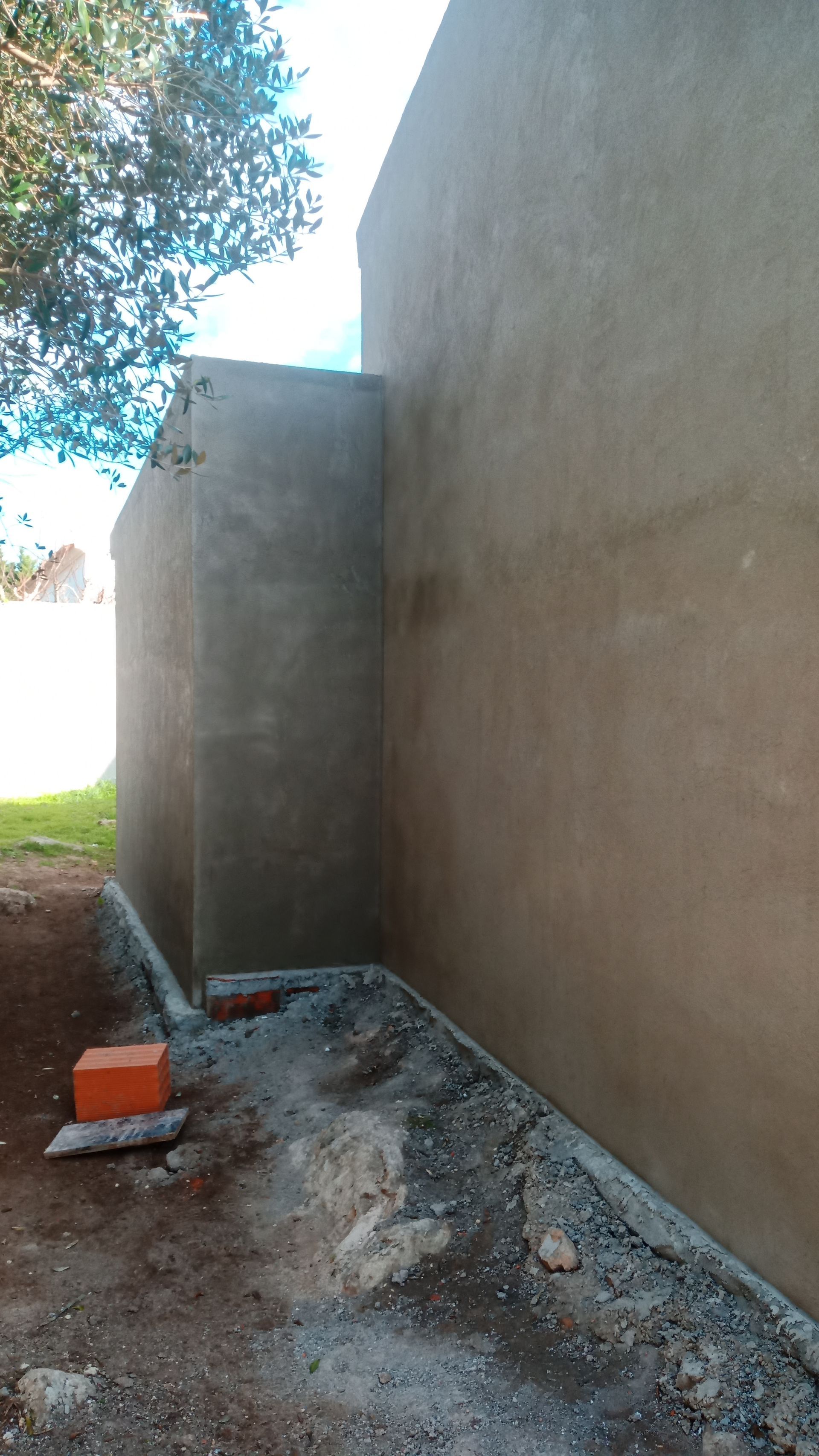 Vista exterior das paredes recém-rebocadas, com tijolos, terra e uma árvore visíveis.