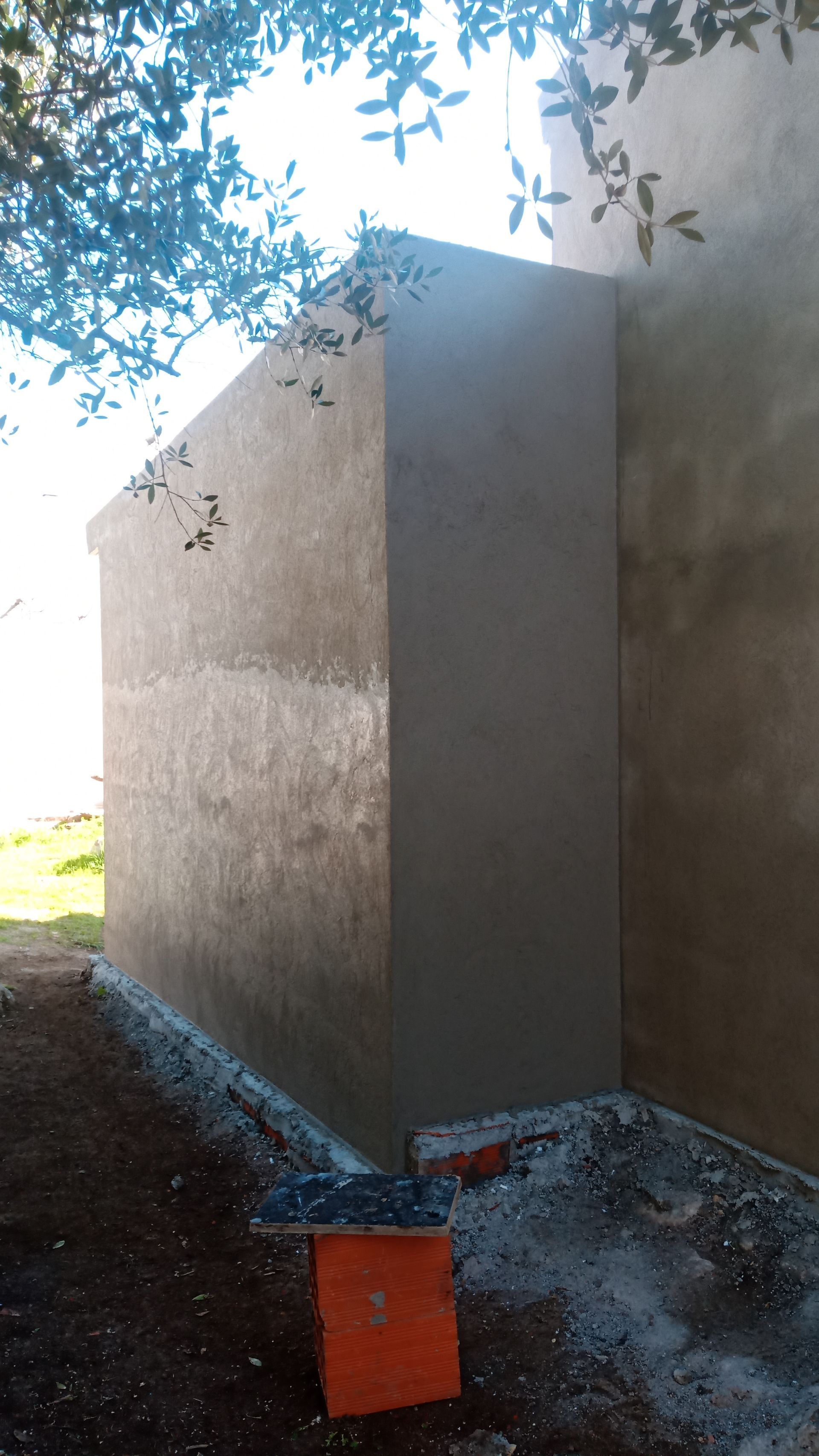 Parede externa de estuque cinza com base de concreto. Uma caixa de ferramentas laranja está na parte inferior. Luz solar filtrada pelos galhos da árvore.
