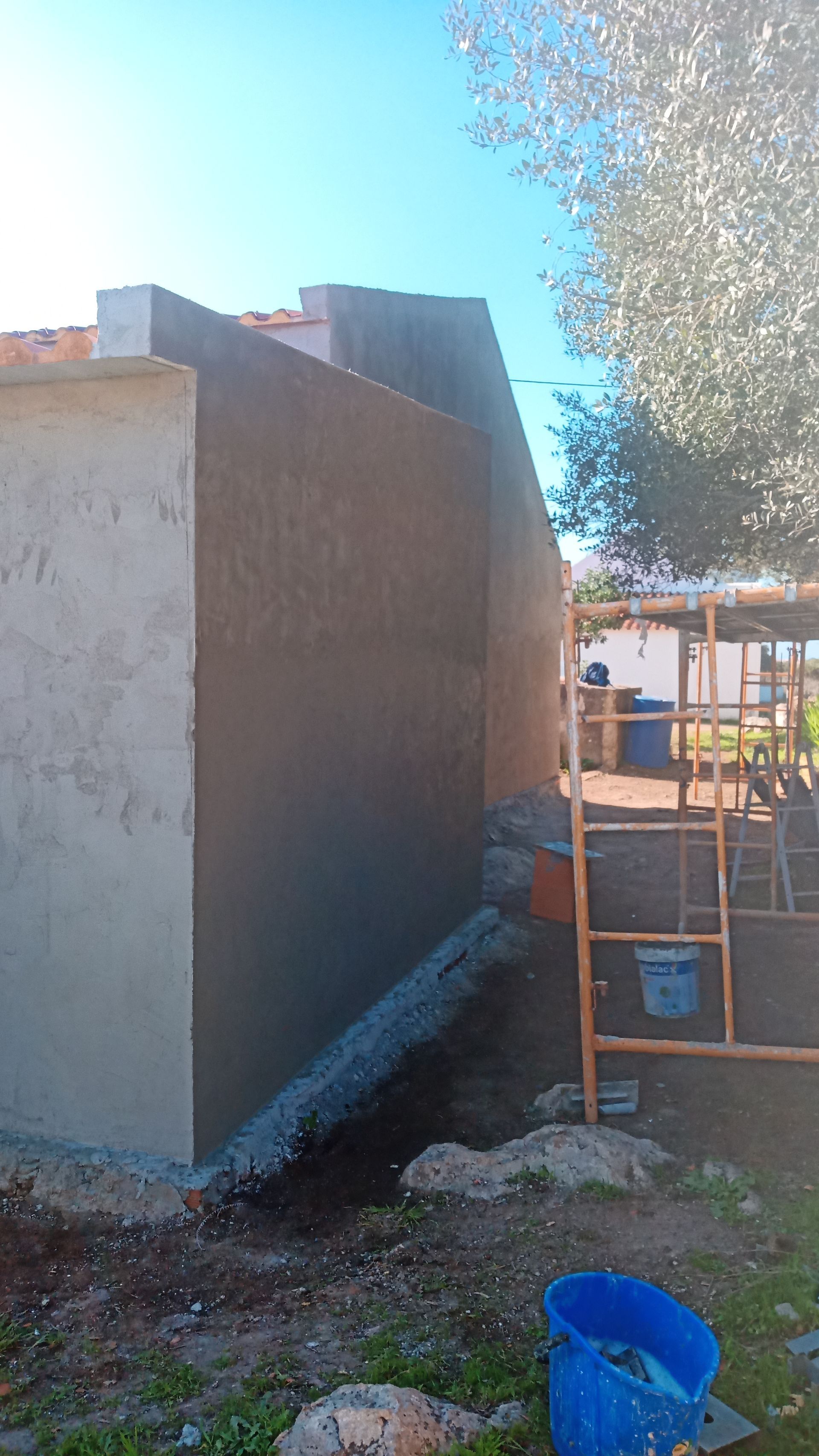Parede externa de um edifício em construção, parcialmente rebocada, com um balde e uma escada azuis próximos.