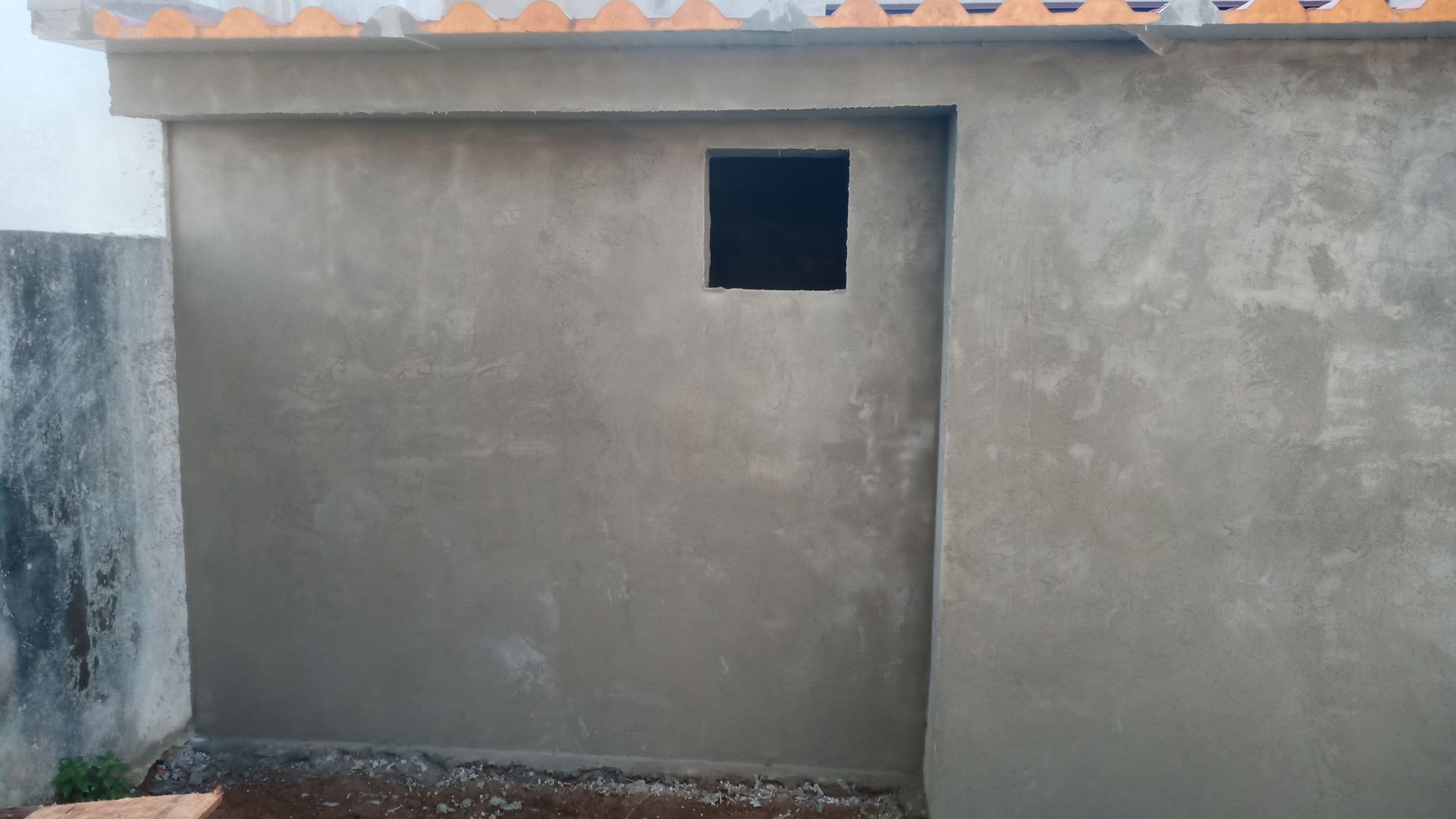 Parede externa rebocada em cinza com uma pequena janela quadrada perto do topo.