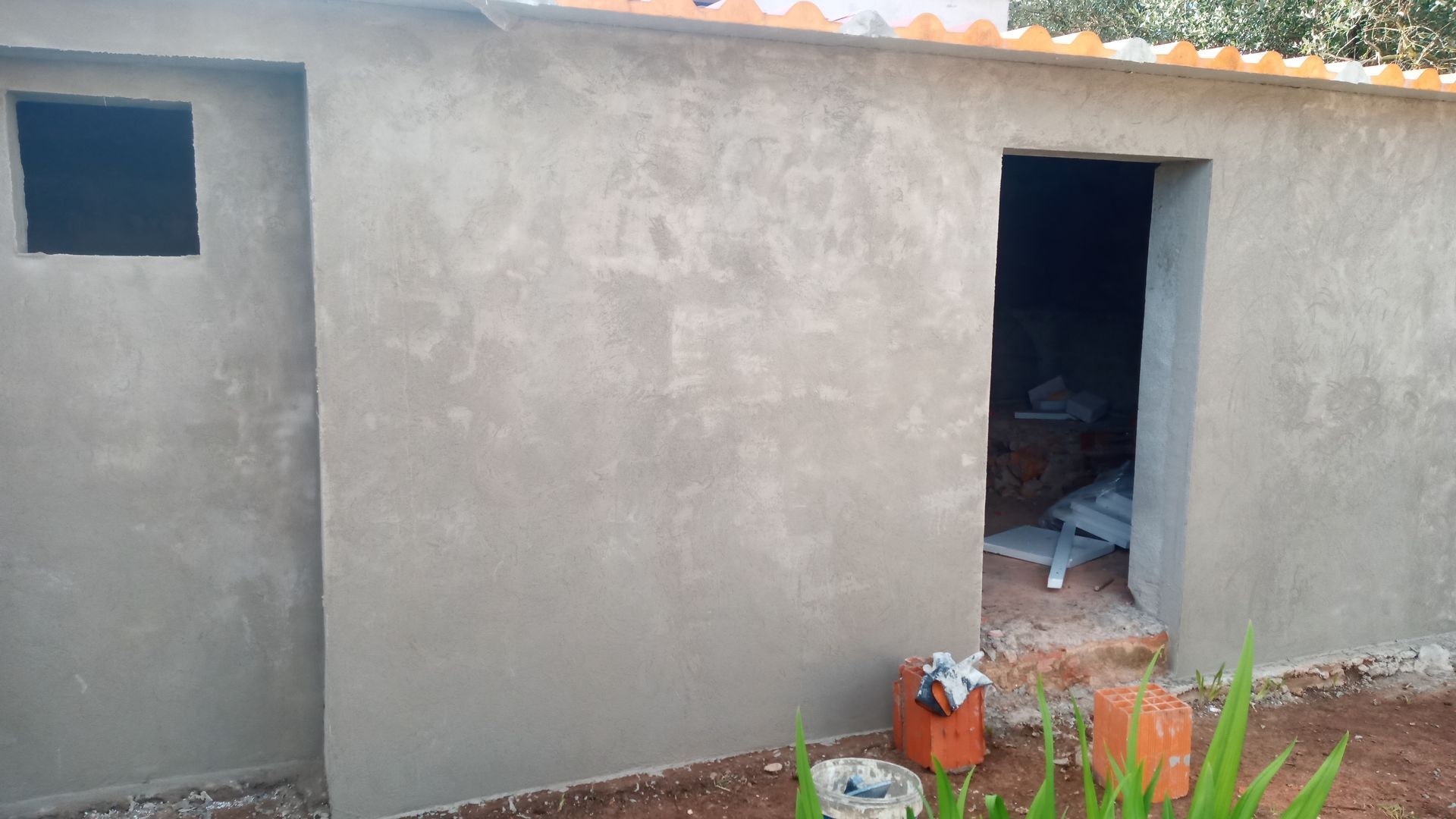 Prédio rebocado de cinza com porta aberta e janela quadrada. Telhas laranja no telhado.