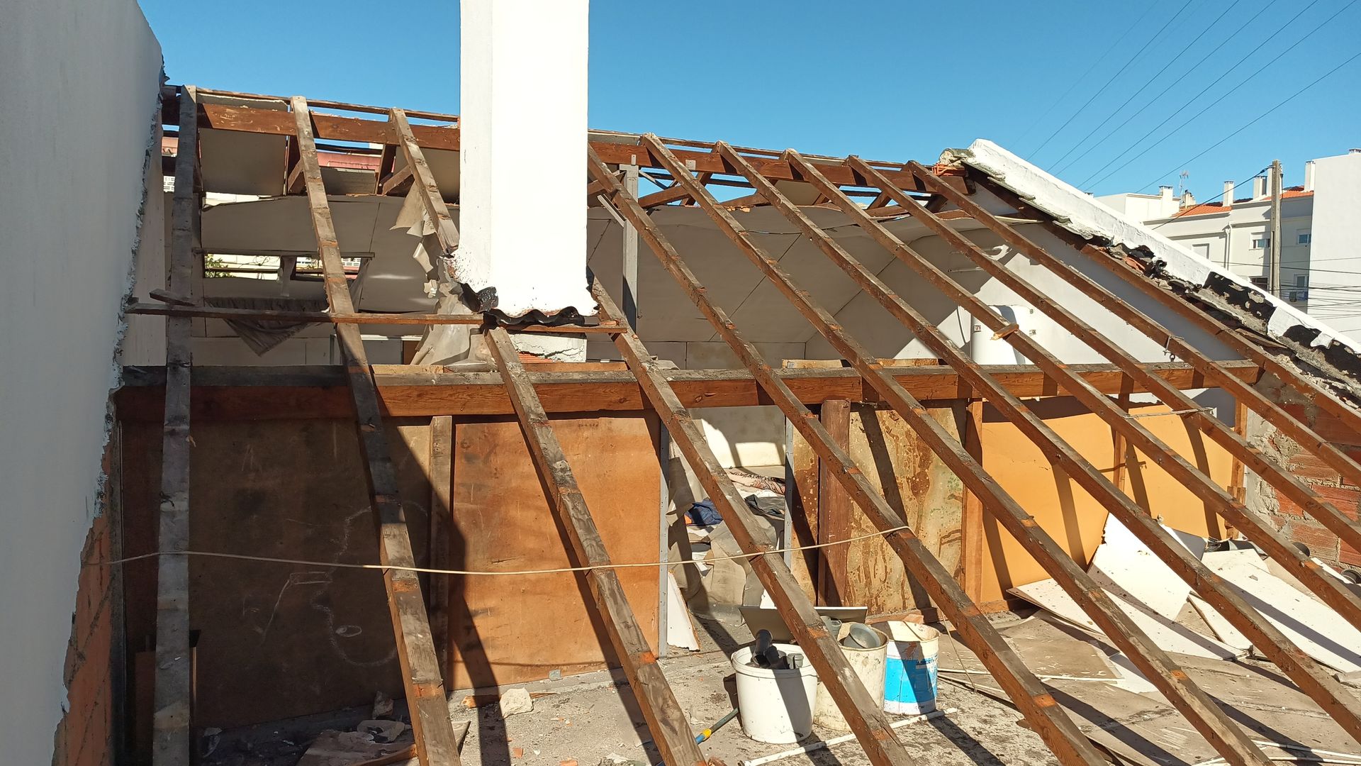 Telhado parcialmente desmontado, mostrando as vigas, caibros e chaminé expostos contra um céu azul claro.