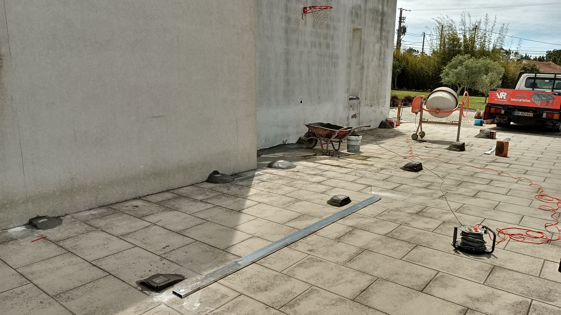 Canteiro de obras com paralelepípedos cinzentos, betoneira e caminhão vermelho perto de uma parede branca.