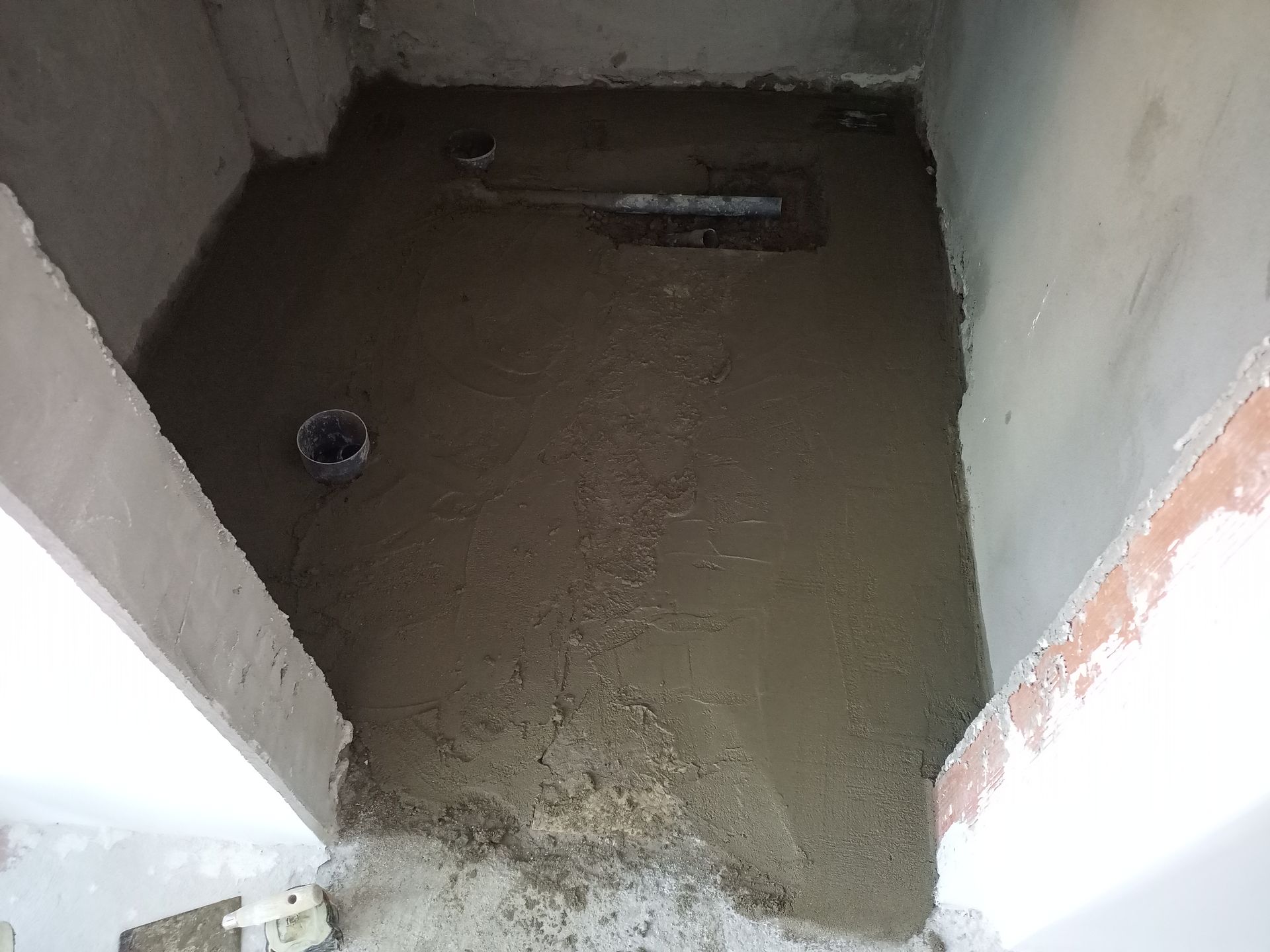 Piso de concreto sendo instalado em um cômodo pequeno e quadrado; instalações hidráulicas visíveis.