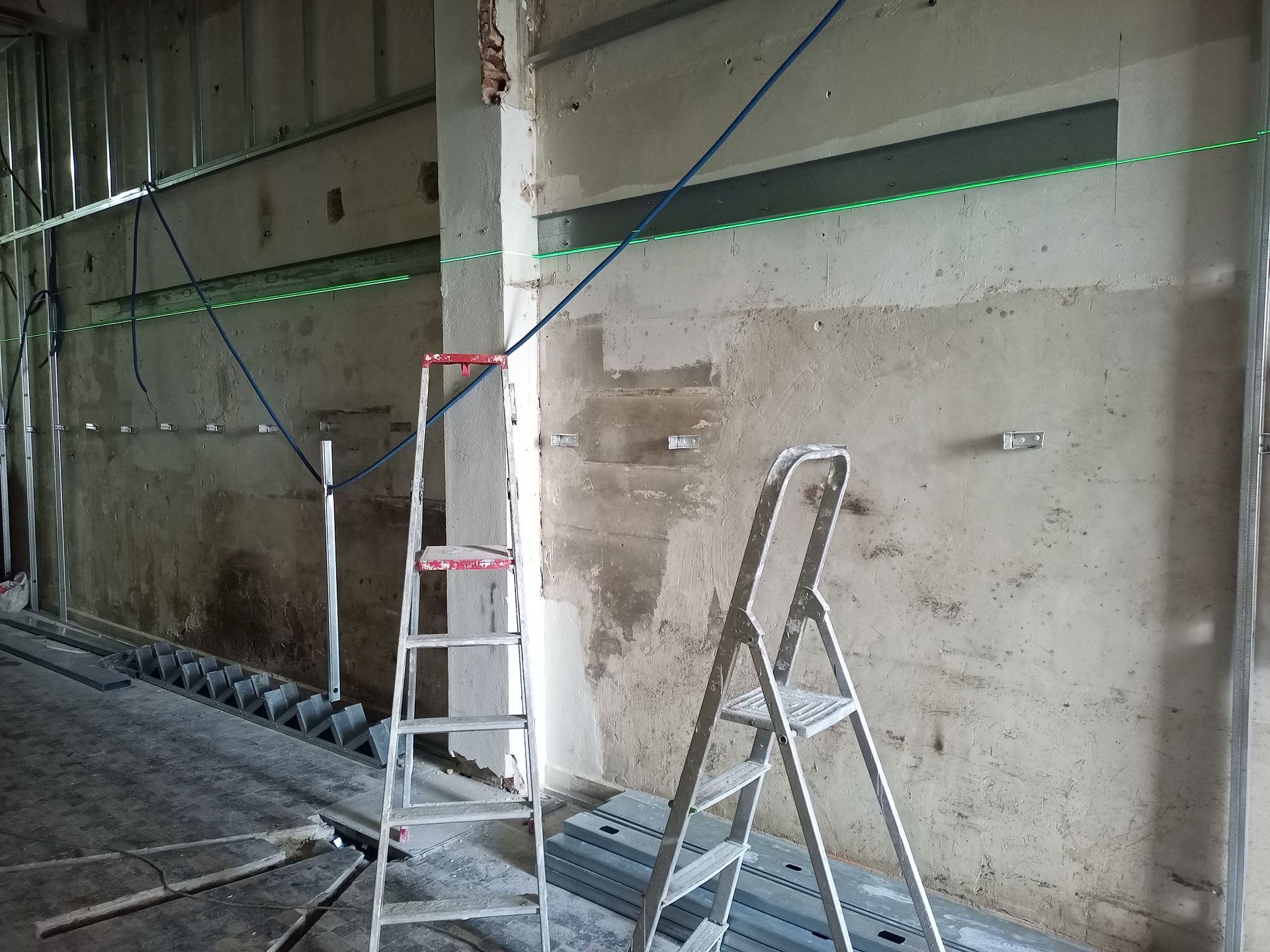 Canteiro de obras interno com escadas, níveis a laser e perfis metálicos. Paredes de concreto aparente.