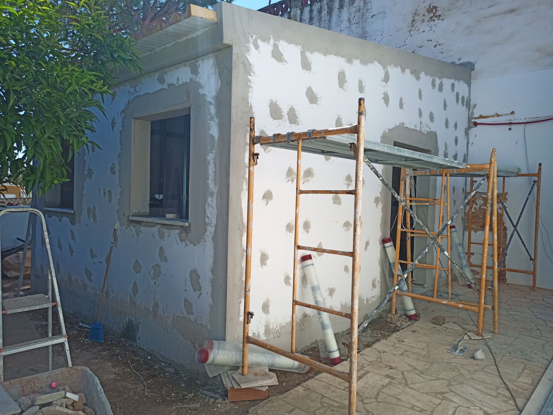 Exterior de um edifício em construção, com andaimes. Paredes cinzentas e brancas, janela e árvore são visíveis.