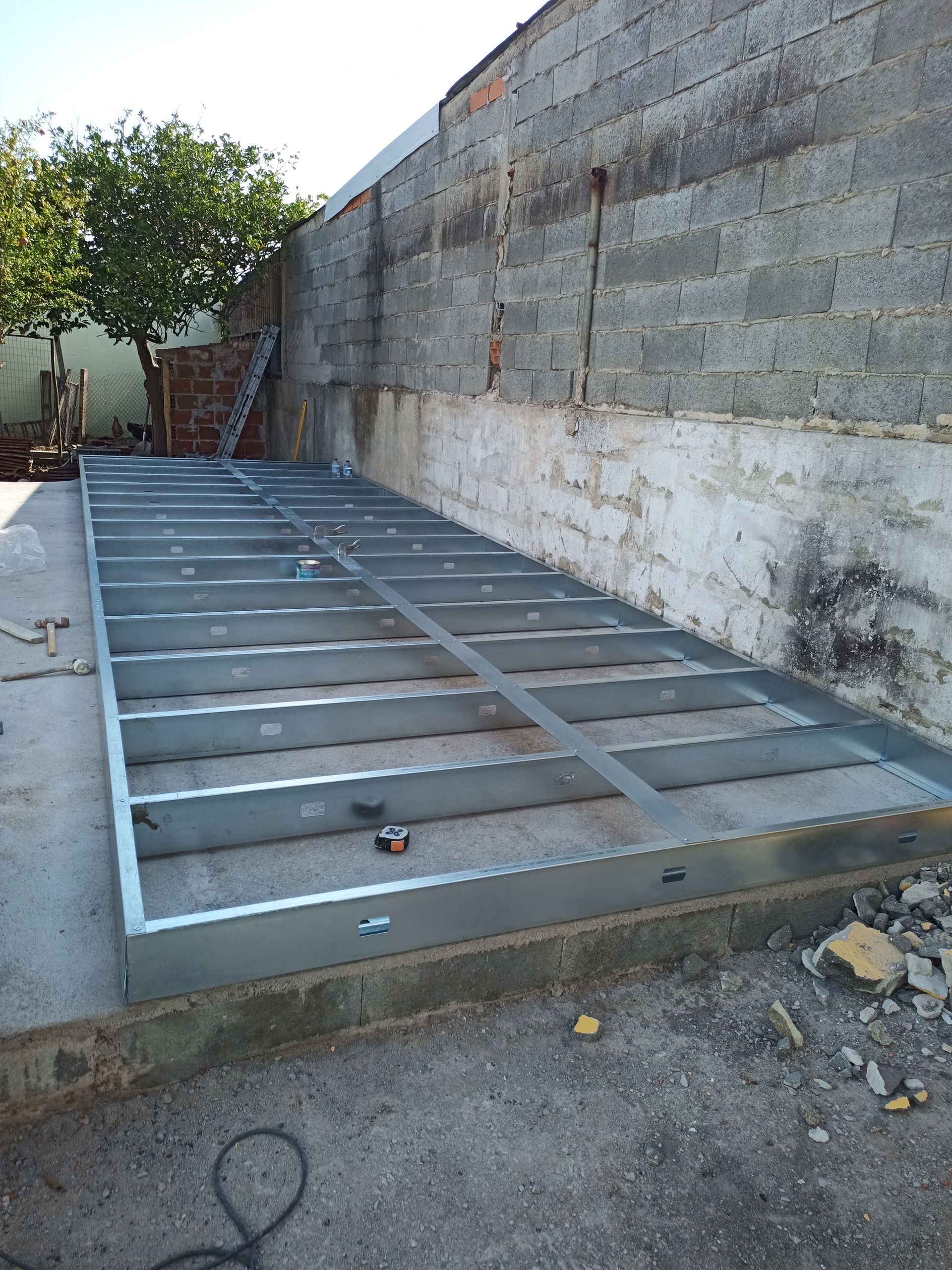 Estrutura metálica assentada sobre concreto ao lado de uma parede de tijolos. Canteiro de obras.