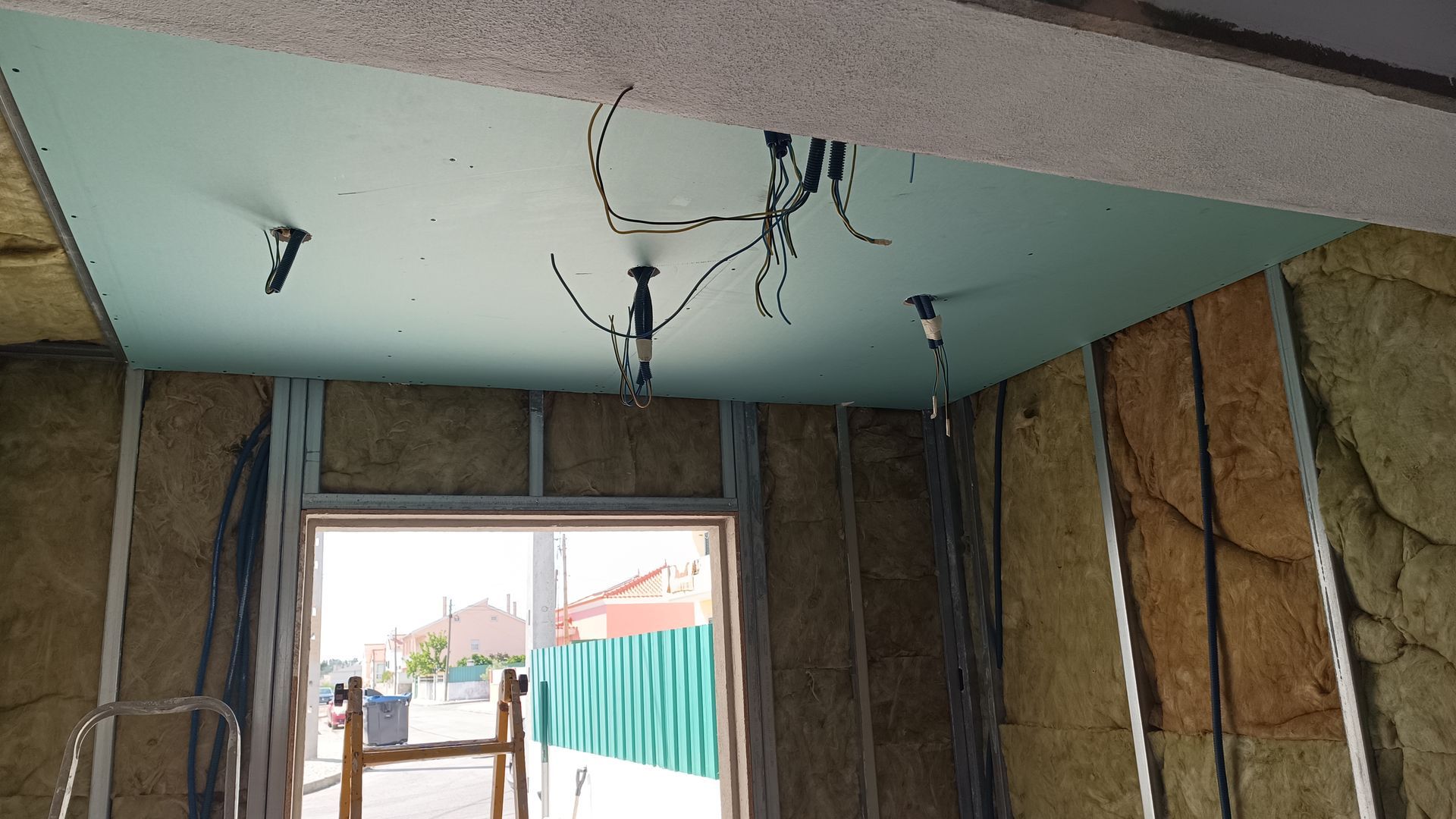 Interior de um canteiro de obras. Paredes e teto isolados com fiação elétrica aparente. Porta aberta.