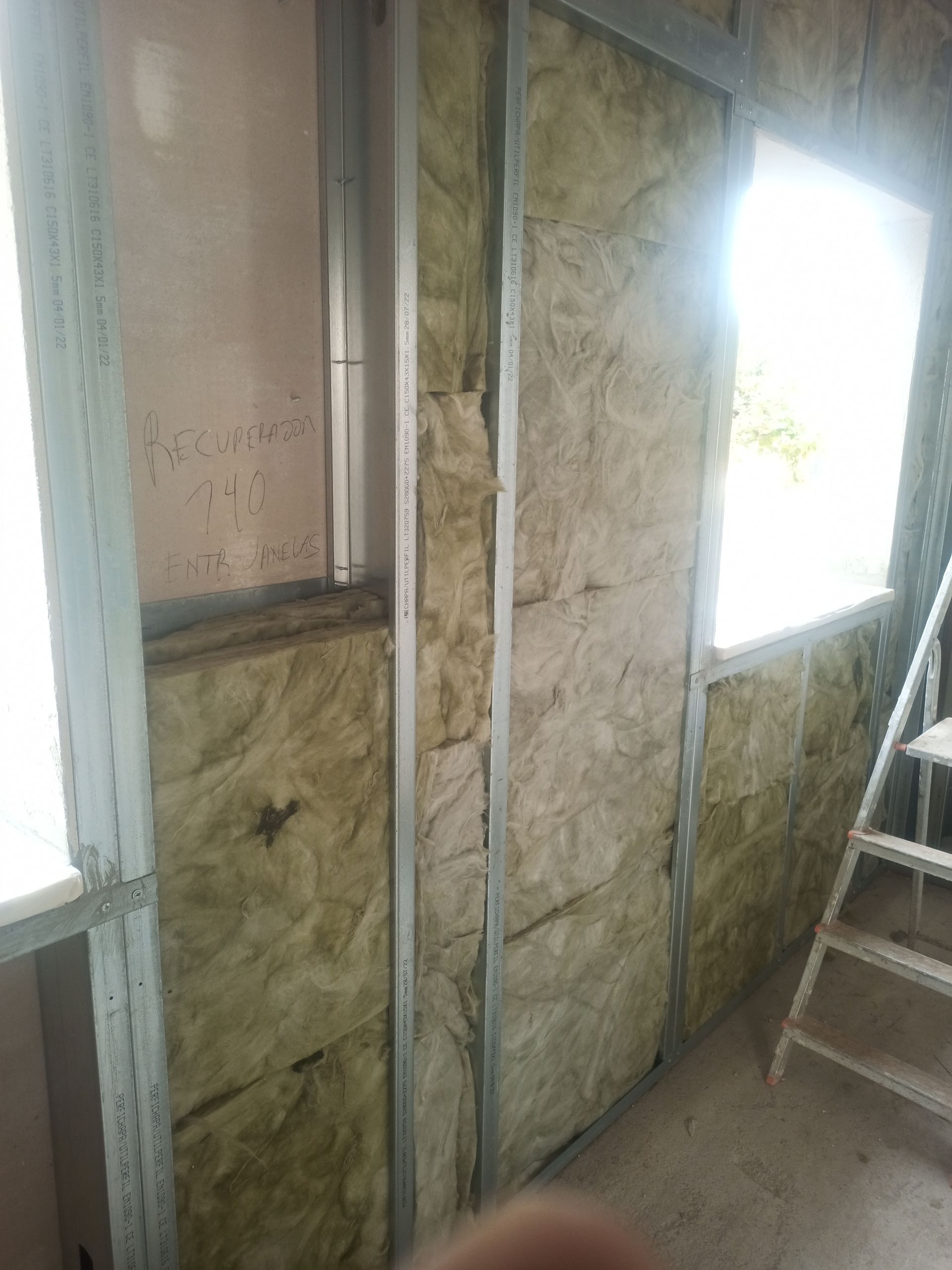 Parede interna em construção com isolamento entre perfis metálicos; janela e escada visíveis.