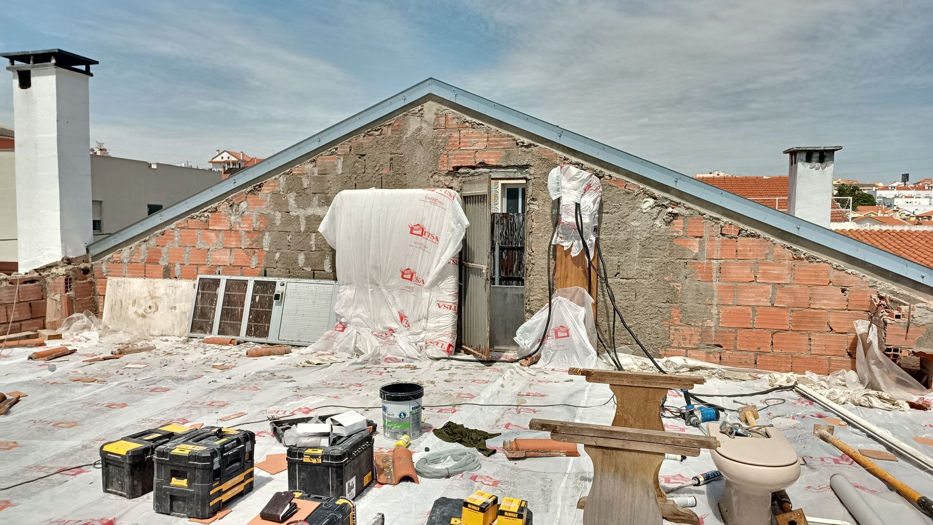 Telhado em reforma com tijolos aparentes, ferramentas e uma vista parcial do interior de um edifício contra um céu claro.