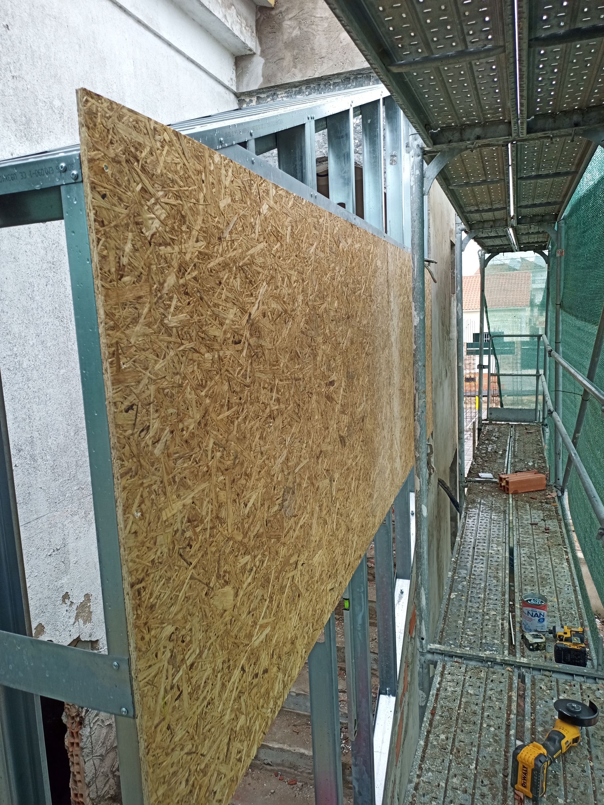 Operário da construção civil instalando placas de OSB na fachada de um prédio com estrutura de metal. Andaimes visíveis.