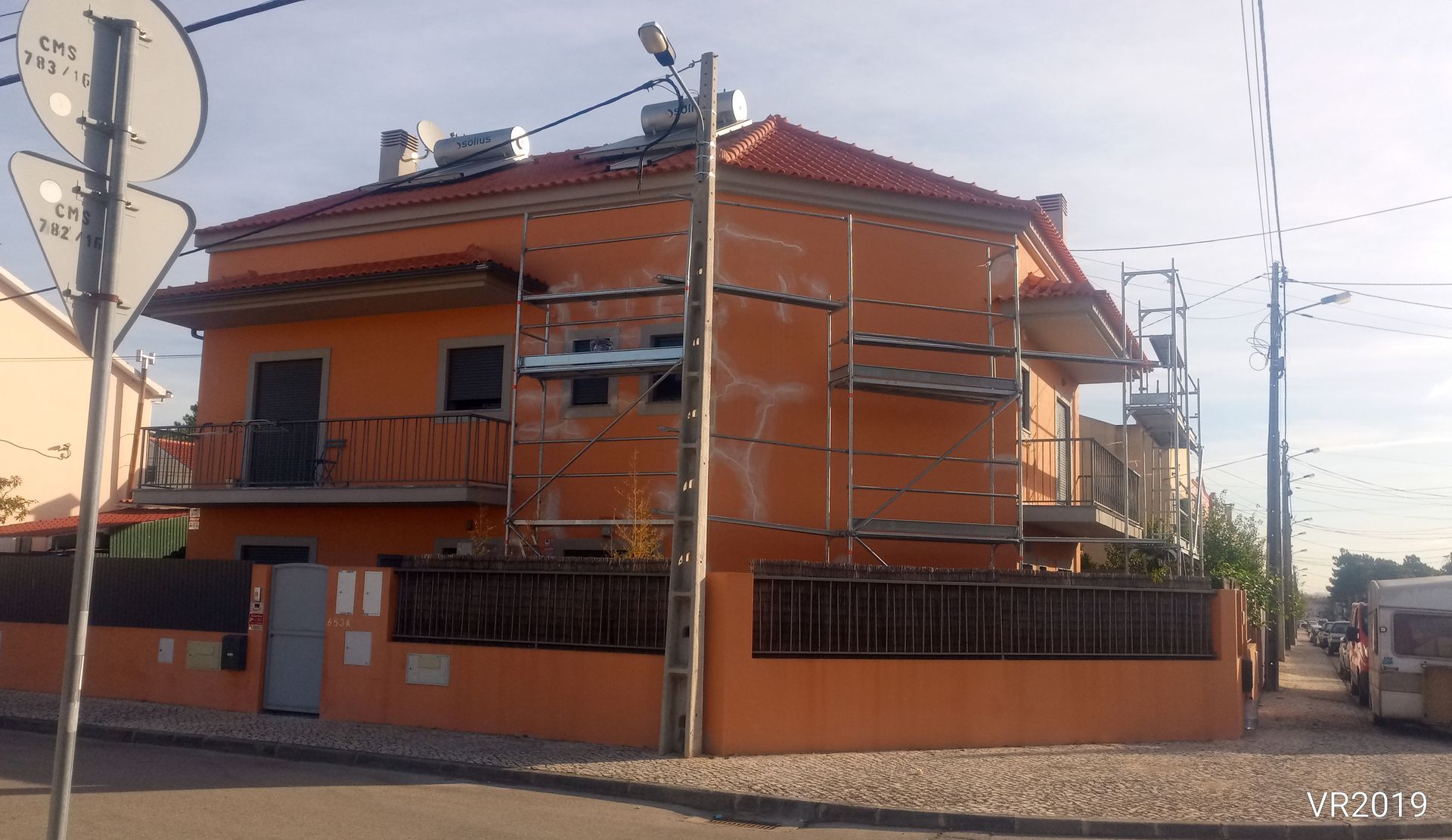 Casa laranja de dois andares com andaimes na lateral, telhado vermelho e cerca baixa.