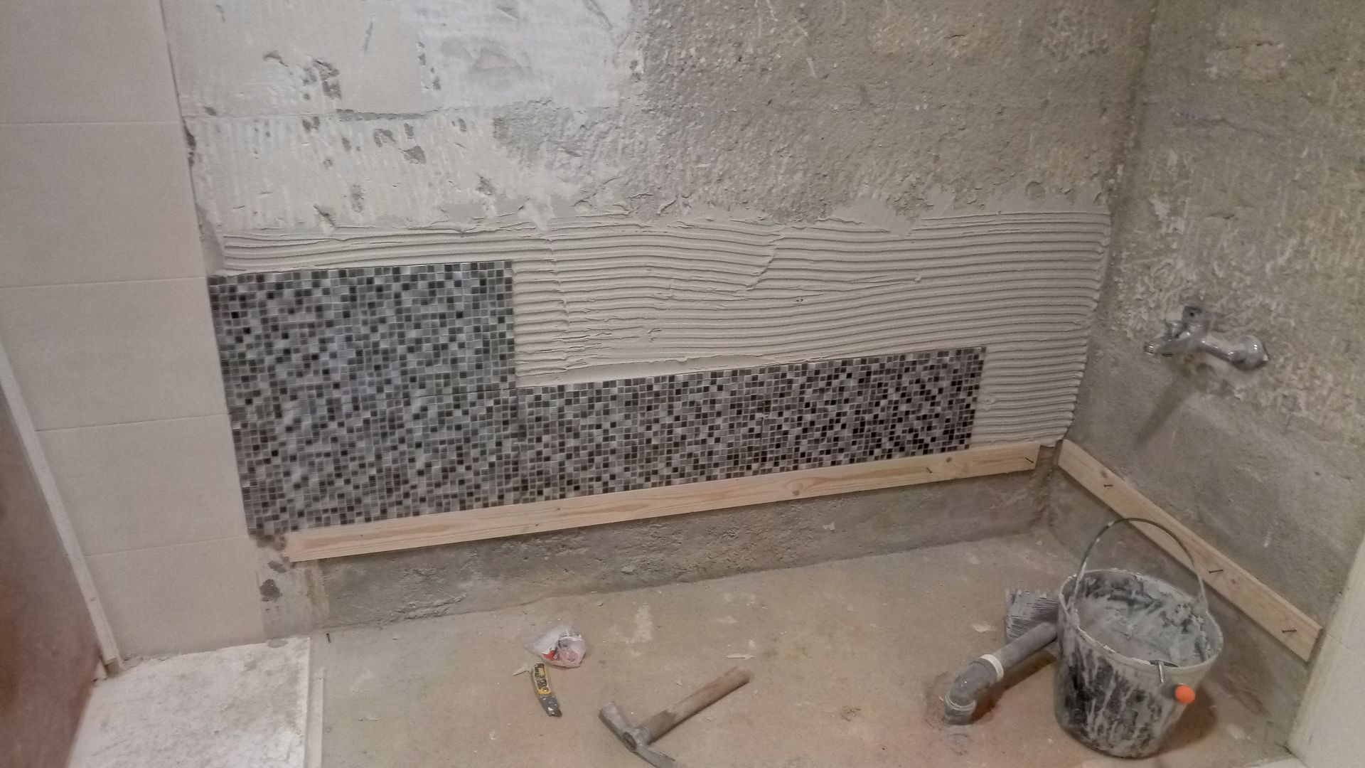 Parede do banheiro parcialmente revestida com azulejos, obra em andamento. Azulejos de mosaico cinza, parede de concreto, balde e ferramentas visíveis.