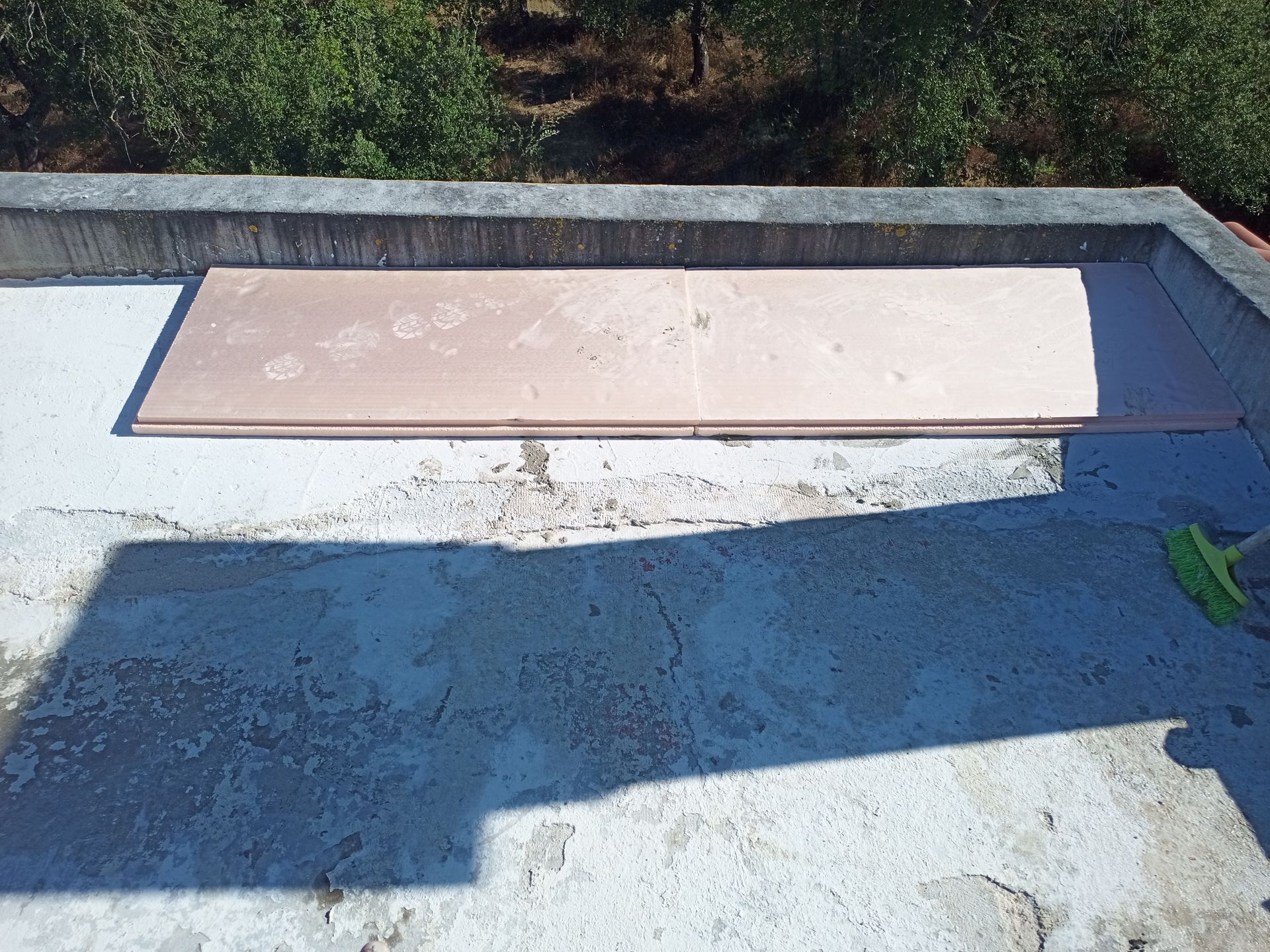 Uma tábua retangular de madeira sobre uma superfície de concreto, com sombras e uma vassoura verde visíveis.