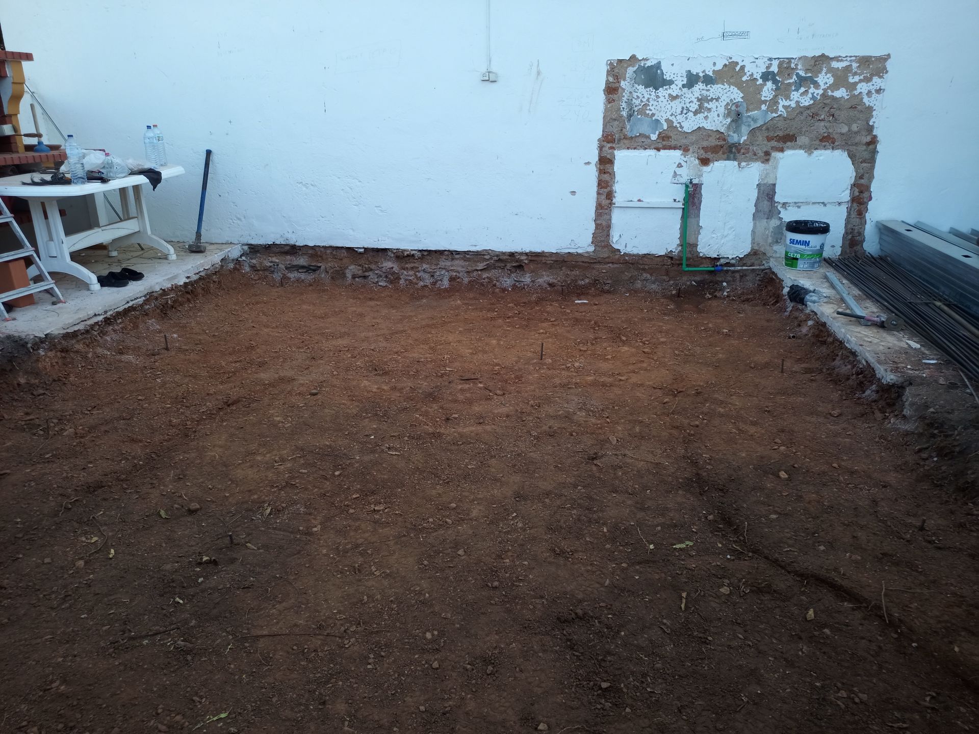 Área de terra escavada junto a uma parede branca, construção em andamento, com entulho e aberturas expostas na parede.