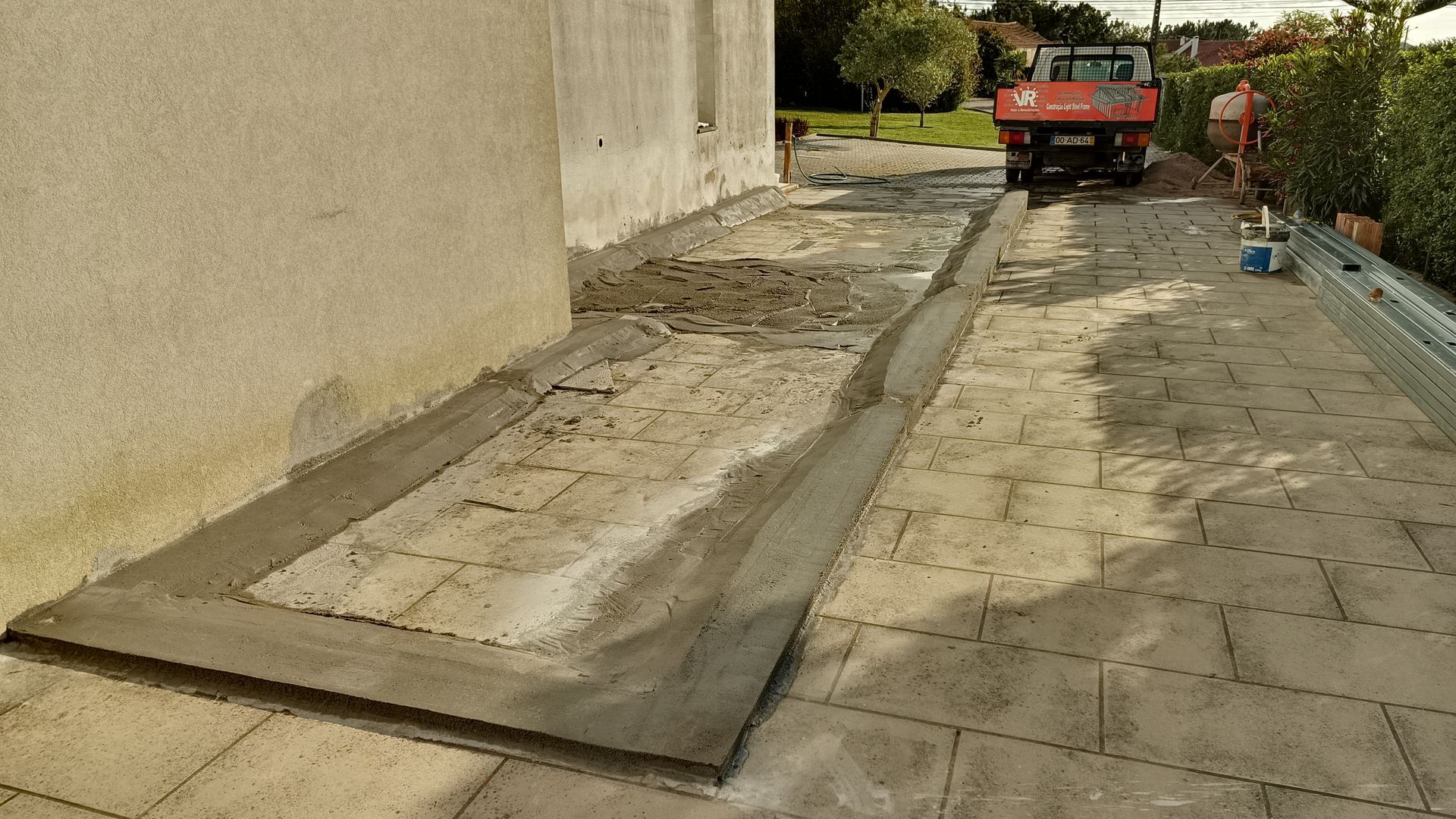 Canteiro de obras: Borda de concreto sendo instalada ao redor de um prédio, ao lado de uma calçada pavimentada. Um caminhão é visível ao fundo.