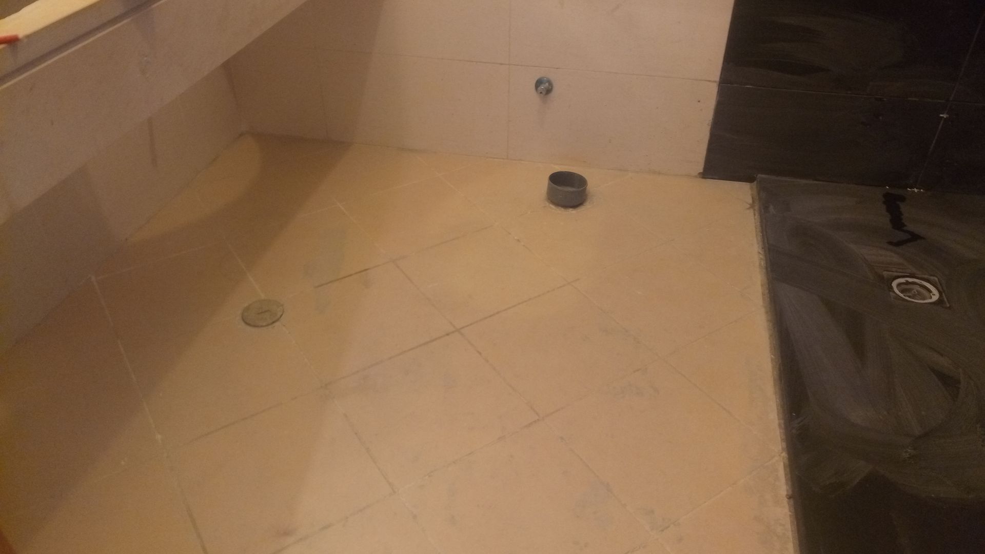 Piso do banheiro com azulejos bege e dois ralos. À direita, há uma área de chuveiro com azulejos pretos e um ralo.