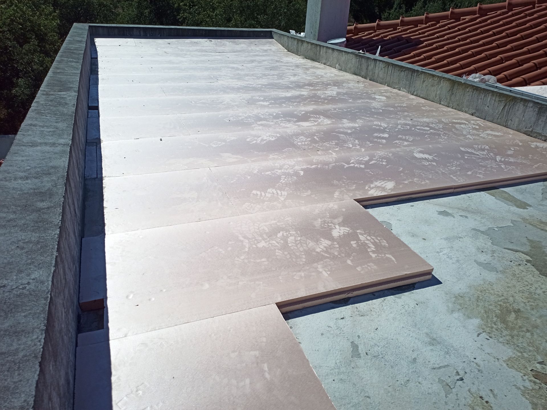 Telhado plano com instalação de painéis isolantes. Perímetro de concreto, telhado de telha ao fundo.