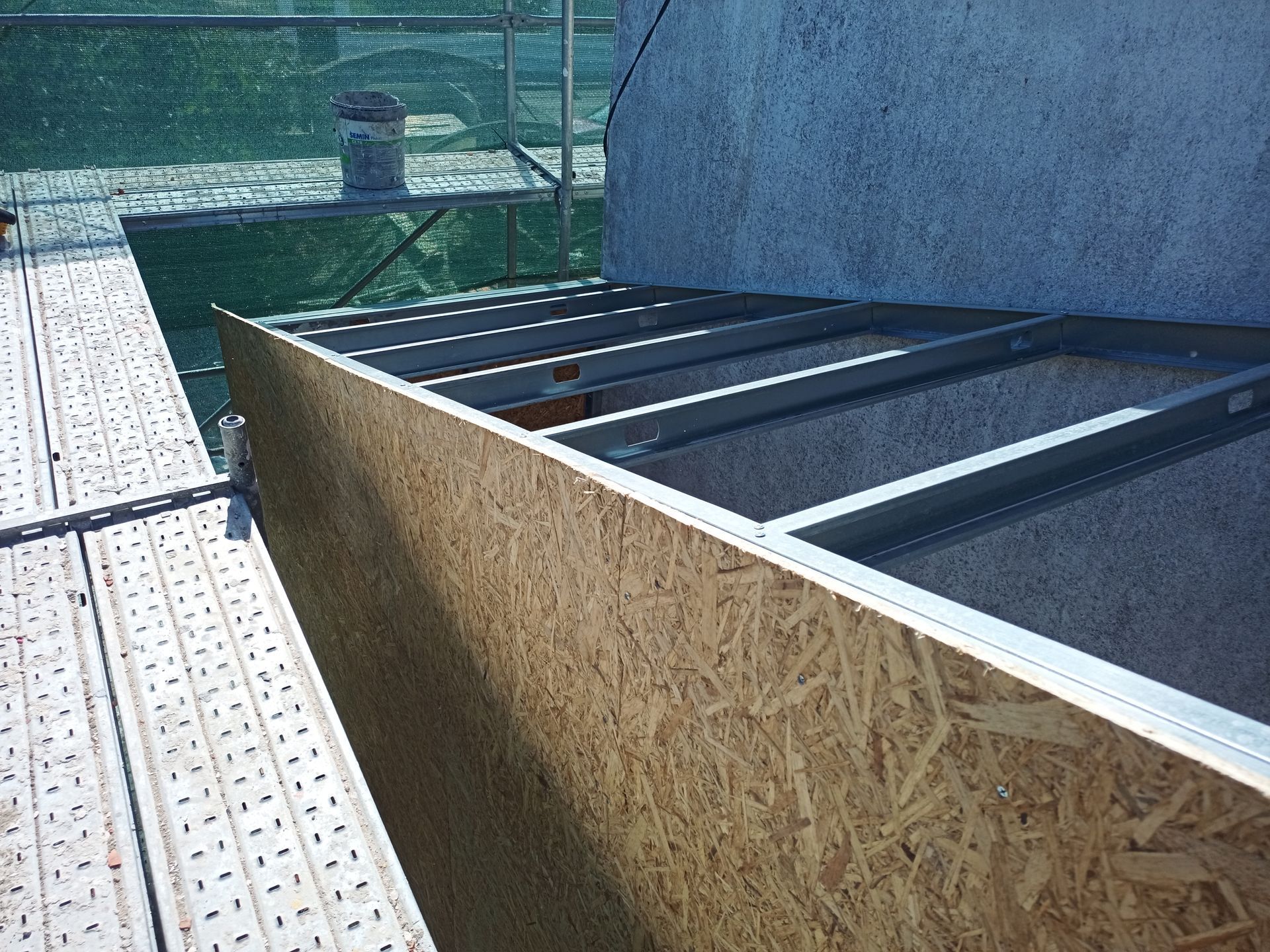 Revestimento de OSB em uma estrutura de telhado, com uma plataforma de andaime em primeiro plano, ao lado de uma parede de concreto.