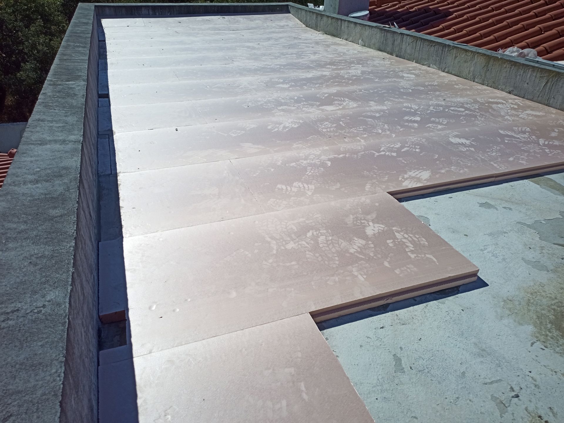 Telhado plano com painéis de isolamento rosa parcialmente instalados. Bordas de concreto e telhado de telha vermelha ao fundo.