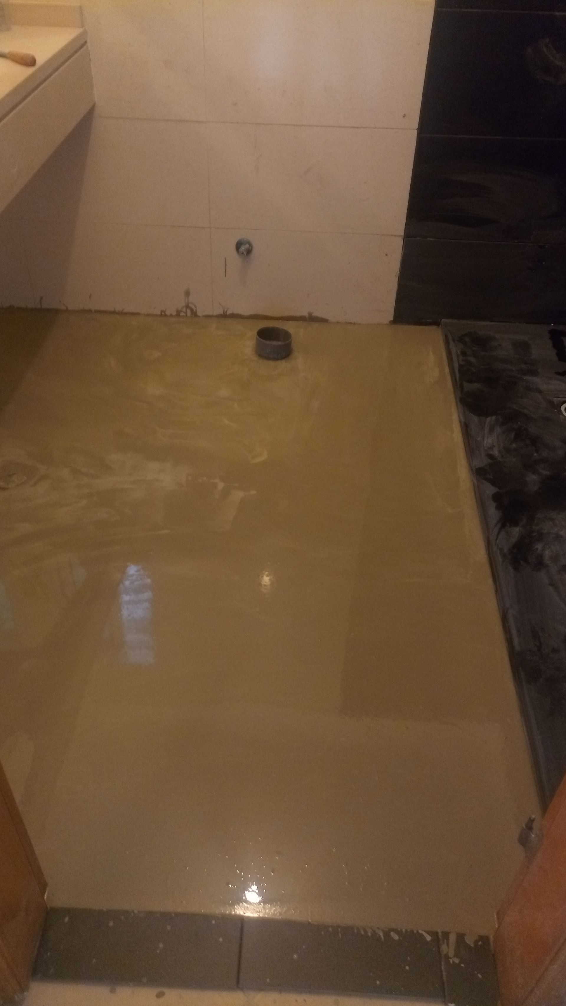 Piso do banheiro, cimento marrom claro, cano de esgoto, borda de azulejo preto, parede branca, armário superior esquerdo.