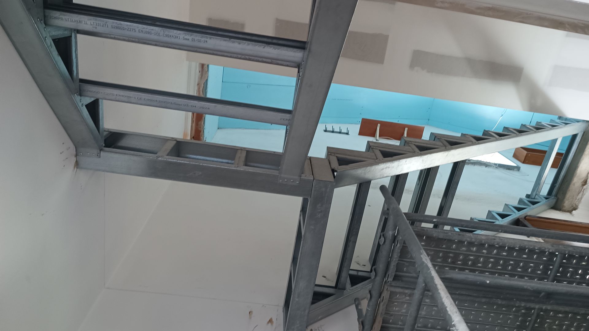 Estrutura metálica da escada, vista de cima. Metal cinza, paredes brancas e vislumbre de azul.