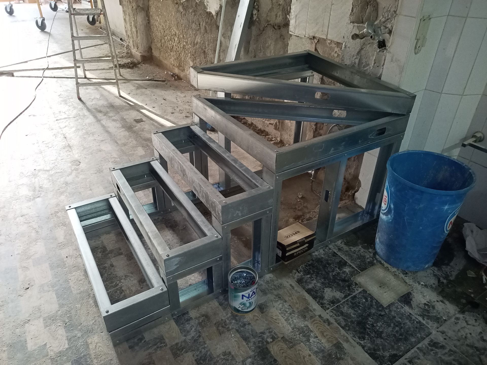 Estrutura metálica para degraus e balcão, em uma sala em construção, com um balde azul e ferramentas.