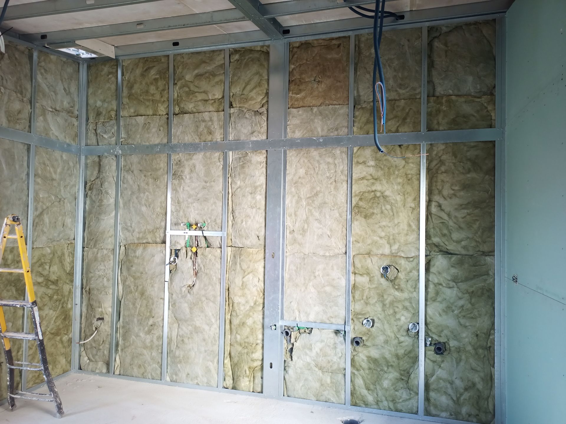 Parede interna em construção com estrutura metálica, isolamento e encanamento aparente.