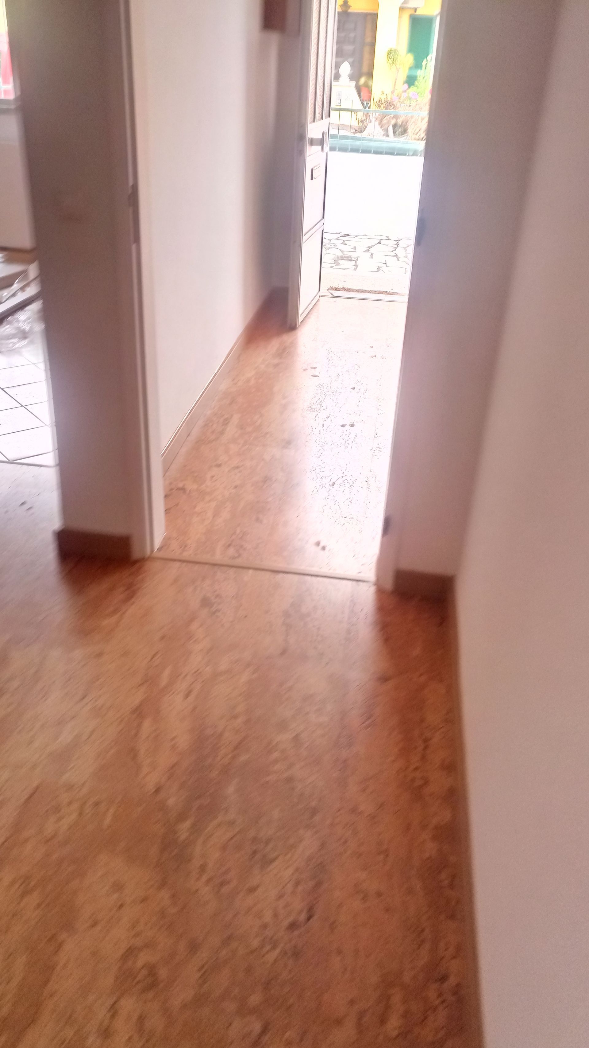 Um corredor com piso marrom, paredes brancas e uma porta aberta que dá para o exterior.